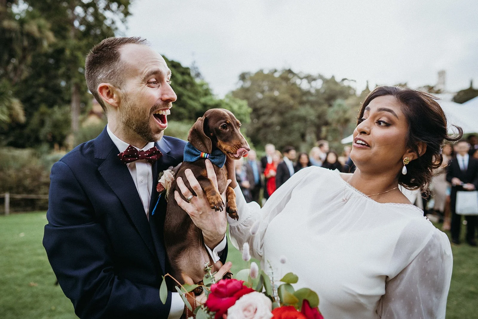 A couple holding their dog who is trying to give them kisses at their wedding