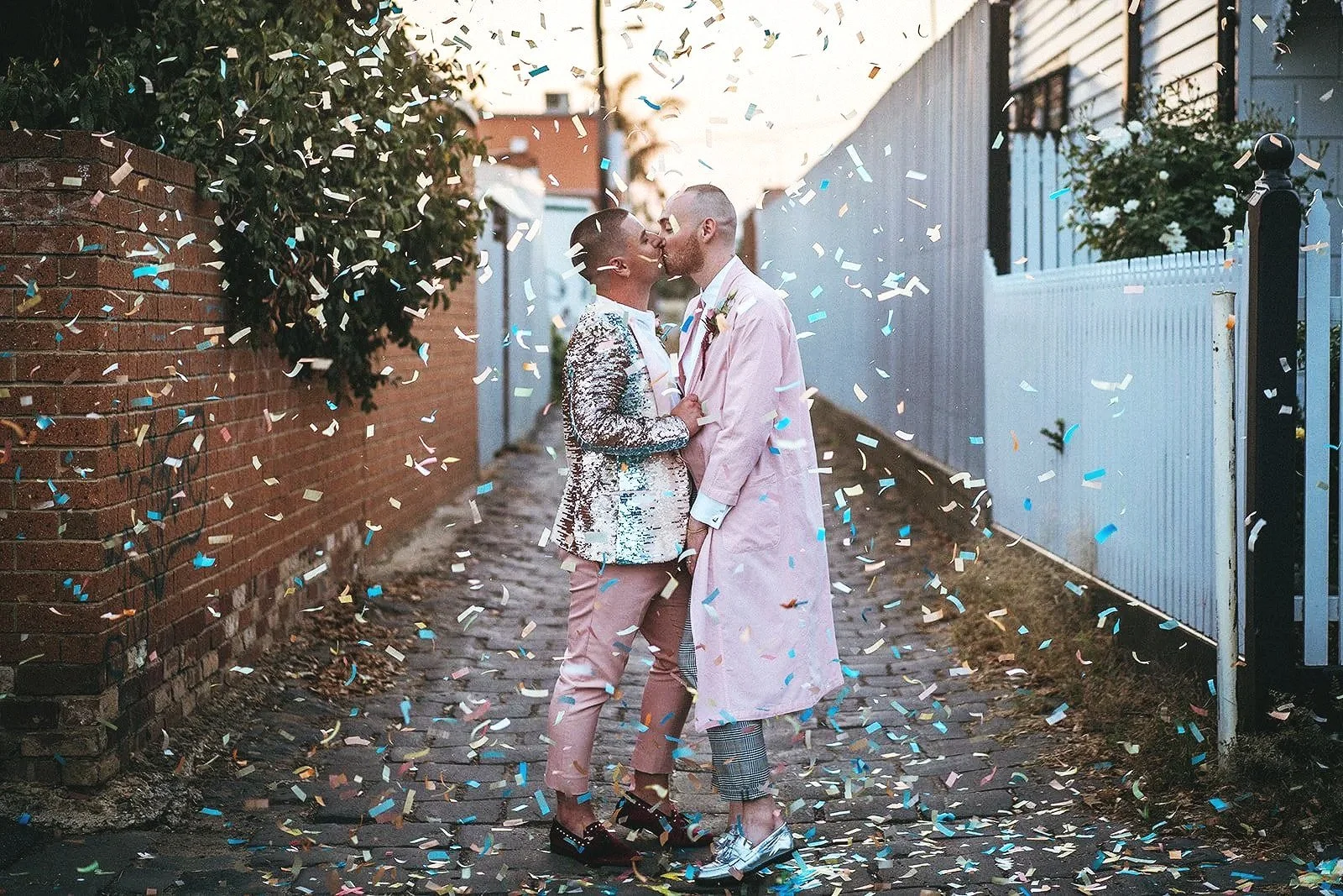 Two grooms dressed in pink kissing beneath falling confetti during colourful Melbourne wedding celebration