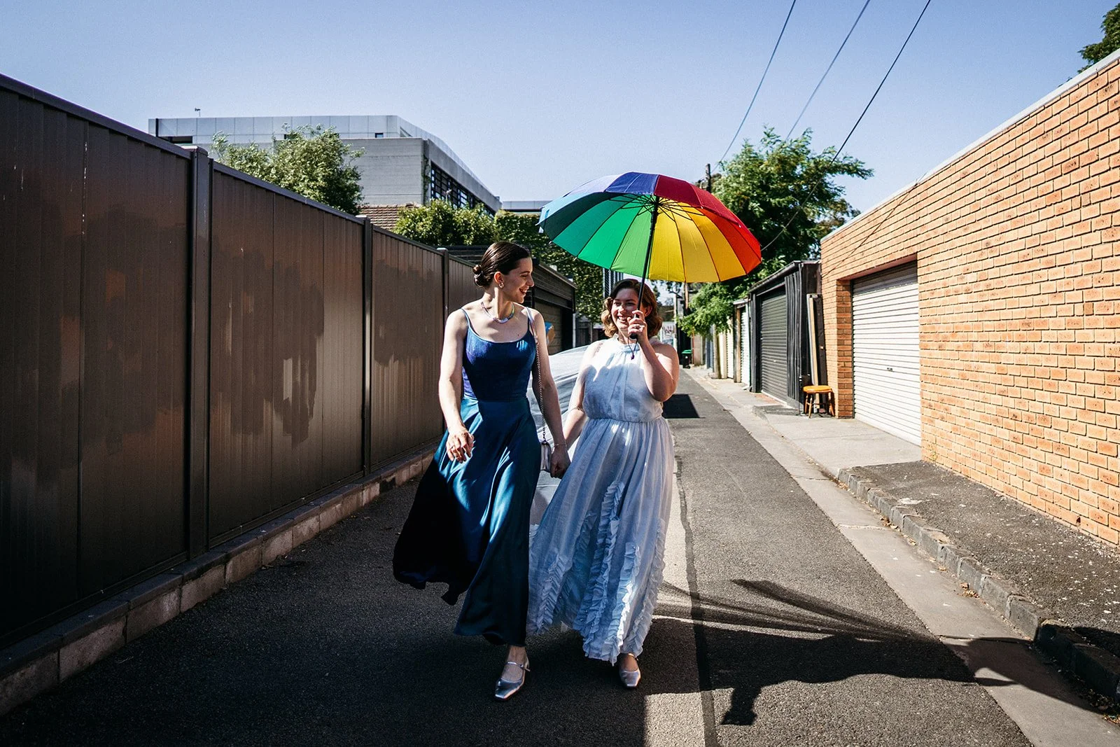 Two Brides walking through Melbourne laneway beneath rainbow pride umbrella