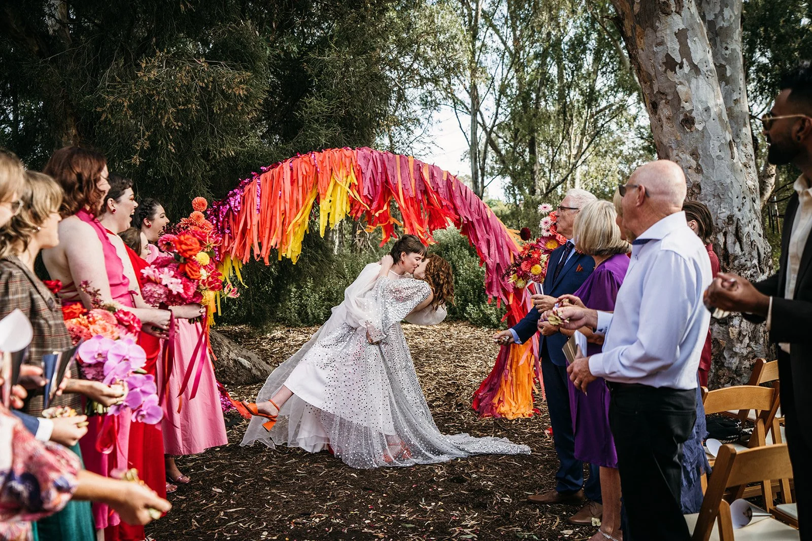 Two brides kissing under a Bangin Hangins colourful arbour during their wedding ceremony at CERES