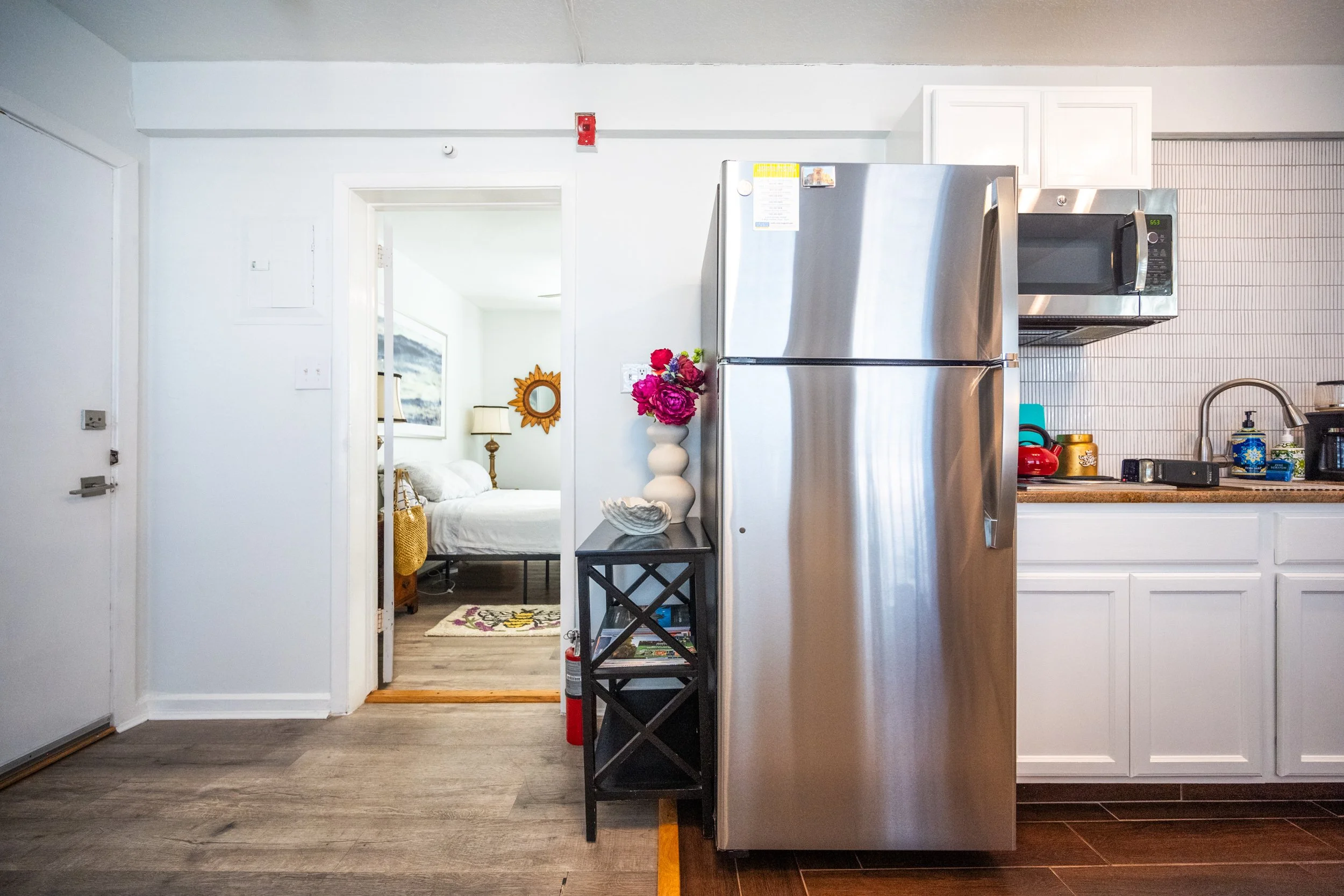 A compact kitchen area with a stainless steel refrigerator, microwave, and sink, with an open doorway leading to a bedroom.