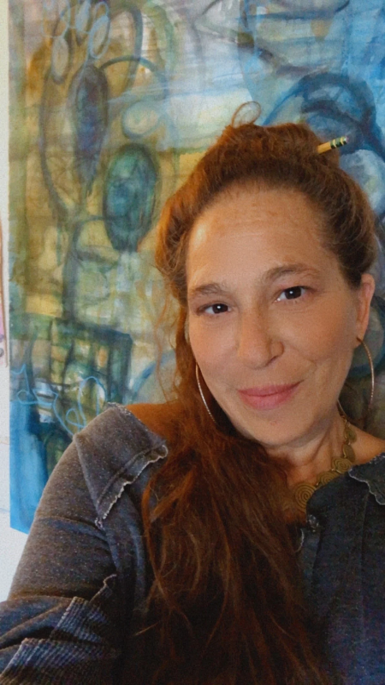 A woman with long red hair, wearing hoop earrings, a necklace, and a dark jacket, smiling at the camera in front of a colorful abstract painting.