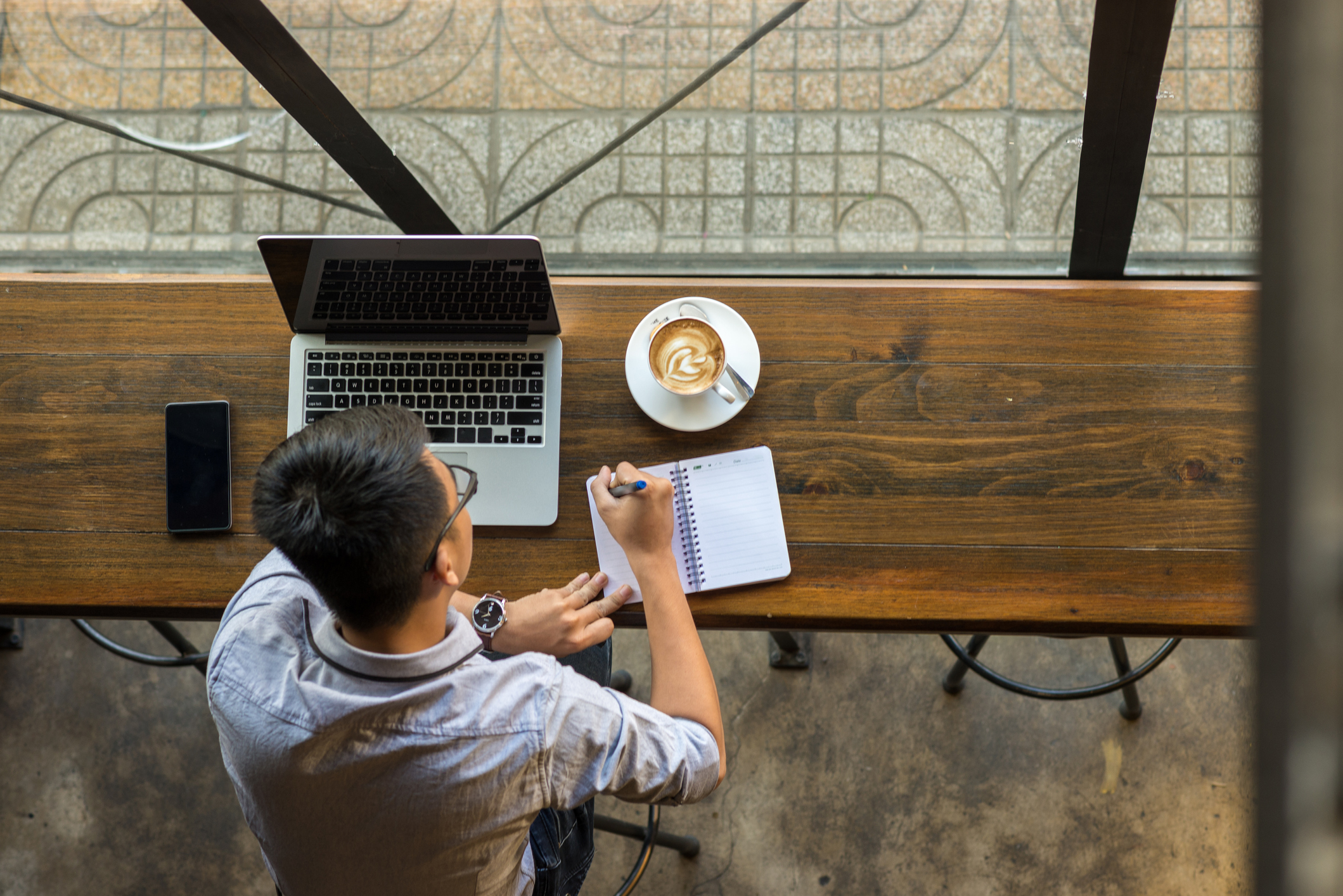 Man writes in a notebook at a wood bar in front of a floor-to-ceiling window.