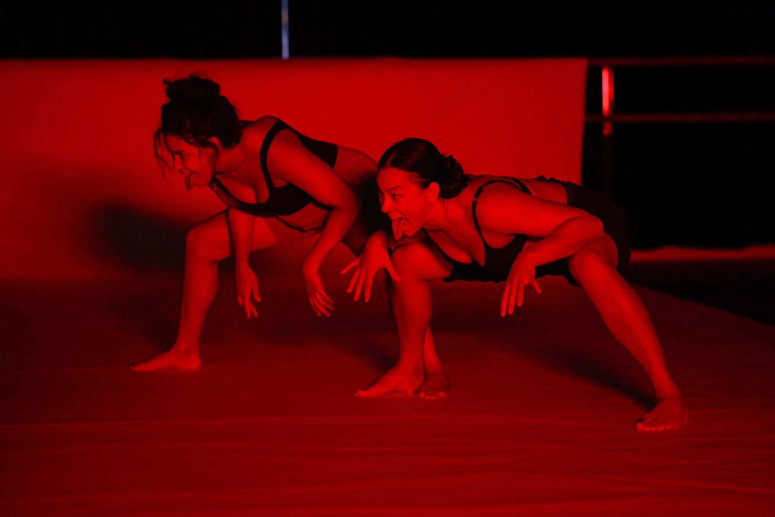 A saturated red photograph of Nora Alami and Kimiko in deep squat. They have active hands and active faces with tongues exposed.