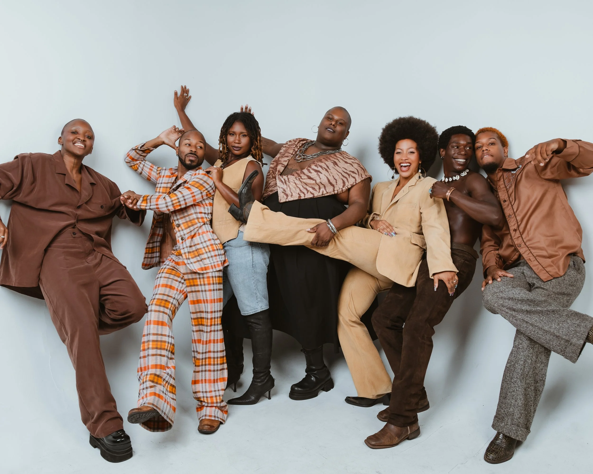 A group of seven Black performers are in coordinating neutral and brown colored outfits, standing against a white backdrop with layered posing.