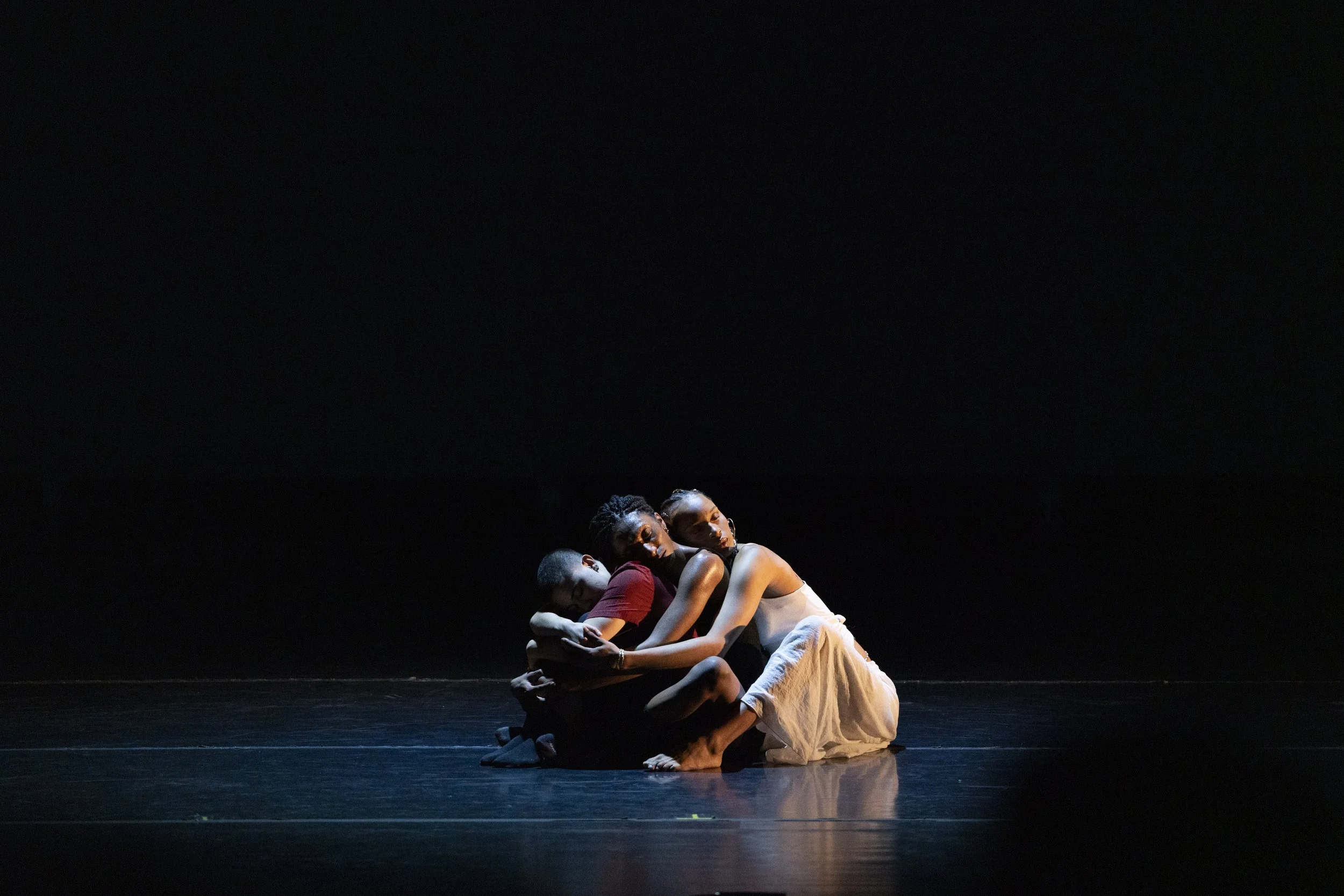 3 trans artists sit in a pool of stage light, holding each other in a shared embrace.