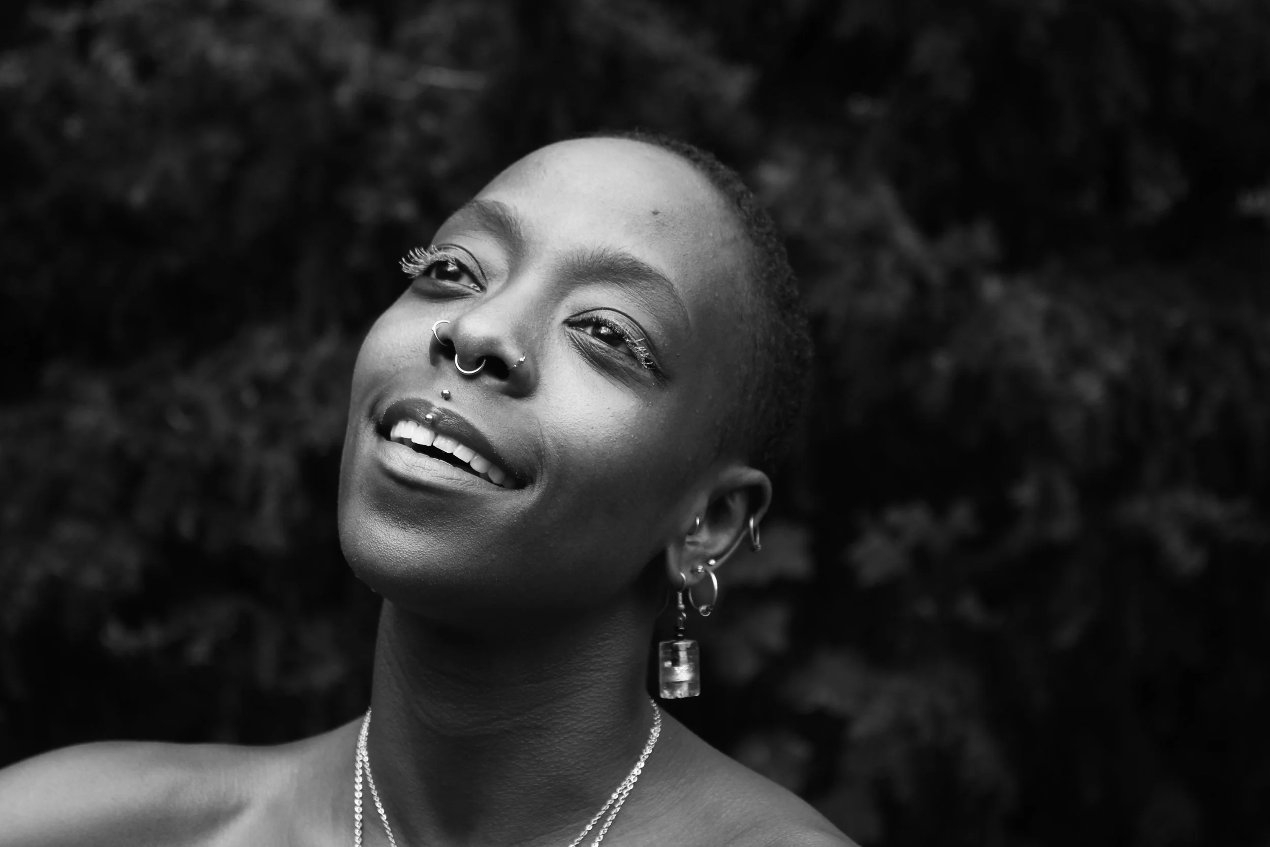 A black and white image of Mawu, a brown skin agender person smiling with their eyes open. Their head slightly tilted.