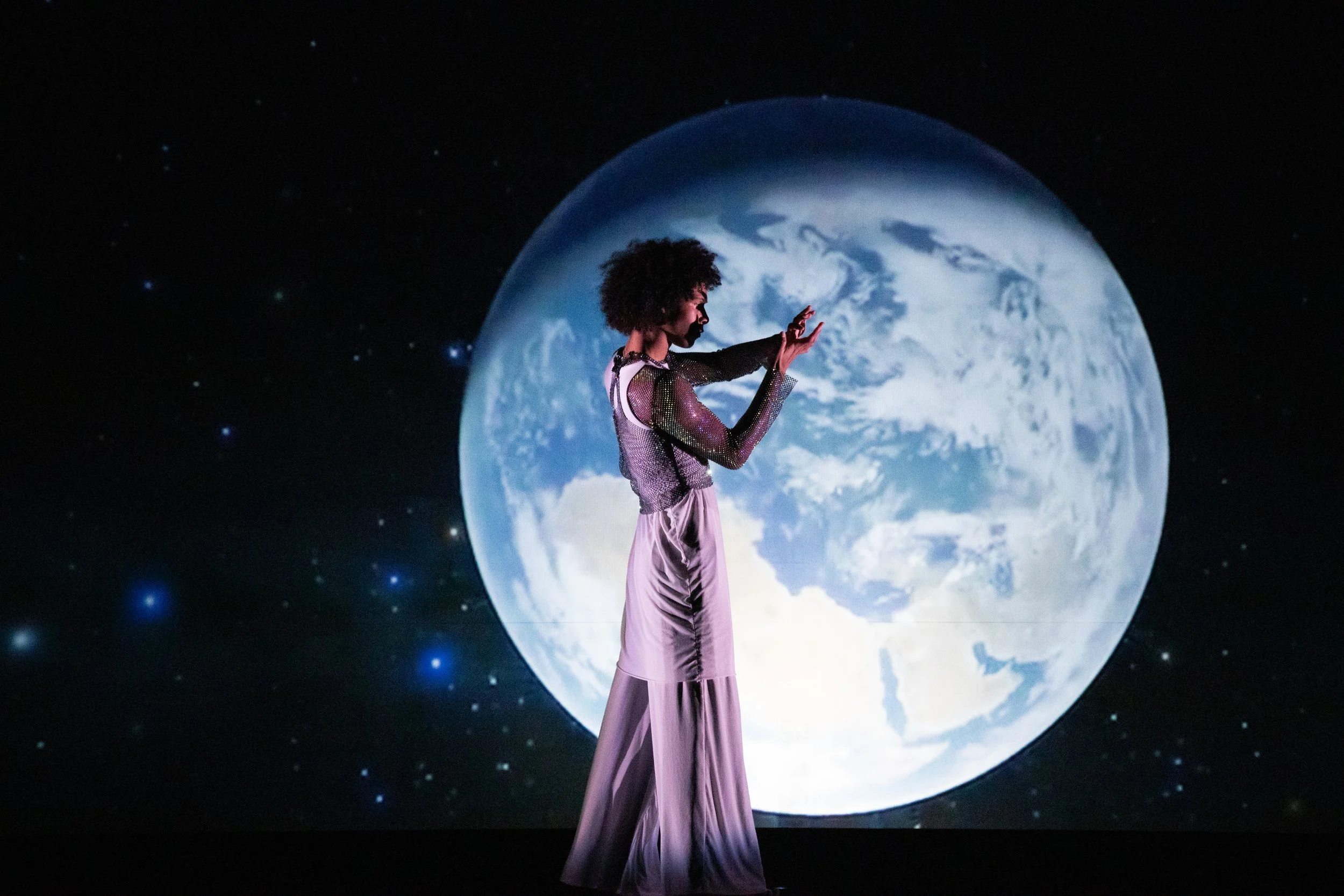 Dylan Richmond stands in a draped white costume investigating the quality of his fingers, silhouetted by planet earth.