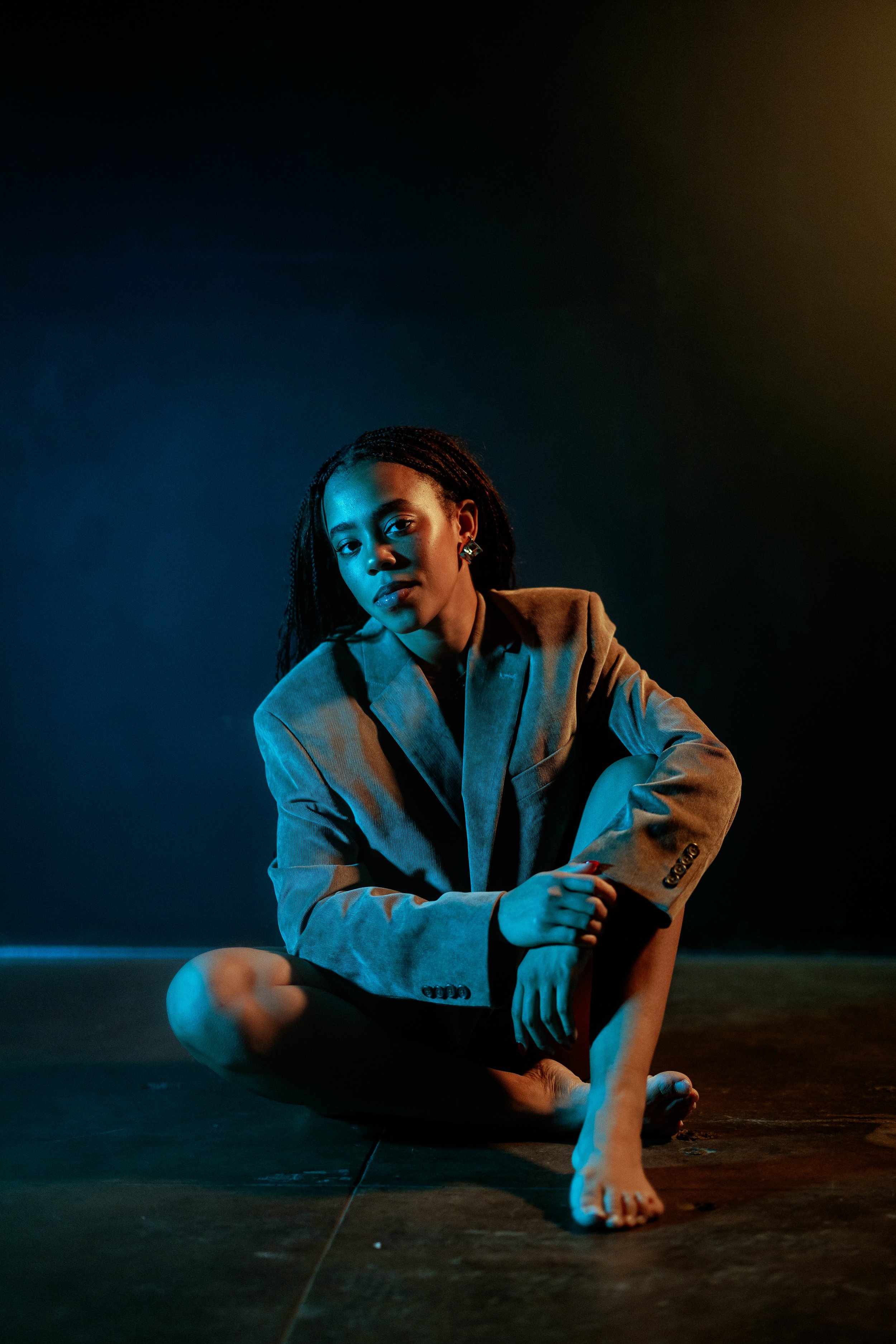 Camryn Stafford seated on the floor in a tan blazer, bare feet, looking directly at camera under blue and amber stage lighting against a dark background.