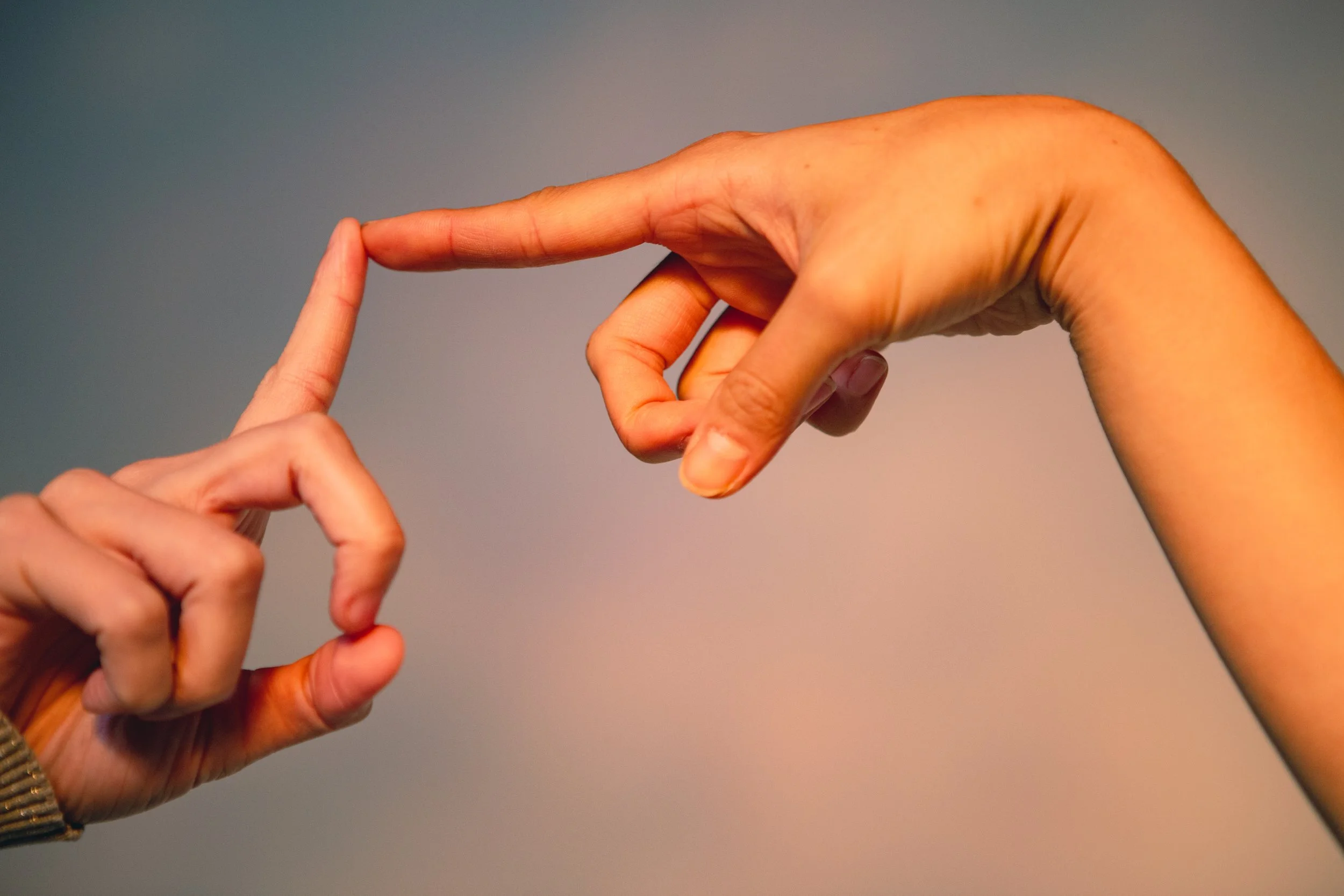 Two different hands, loosely clasped with pointer fingers touching. One hand is higher, wrist limp, one hand is lower, finger erect. Is it sexual? It might be sexual. Background is flat and blue.
