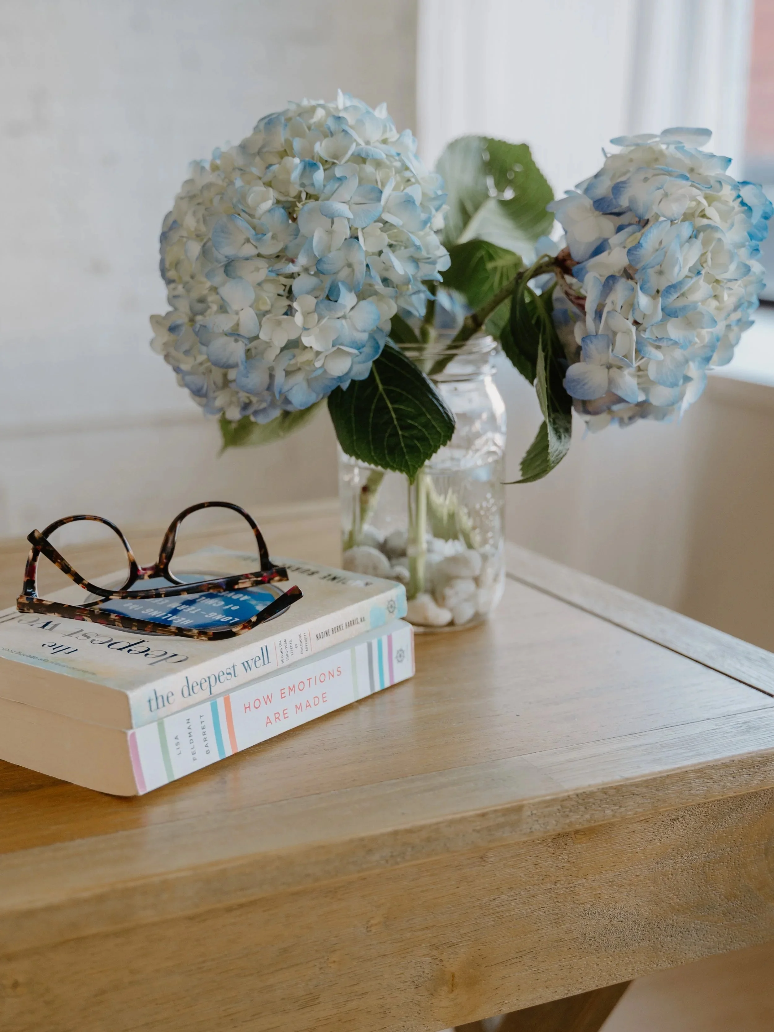 A wooden table with a glass jar holding blue and white hydrangeas, a pair of tortoiseshell glasses, and two books titled 'The Deepest Well' and 'How Emotions Are Made'.