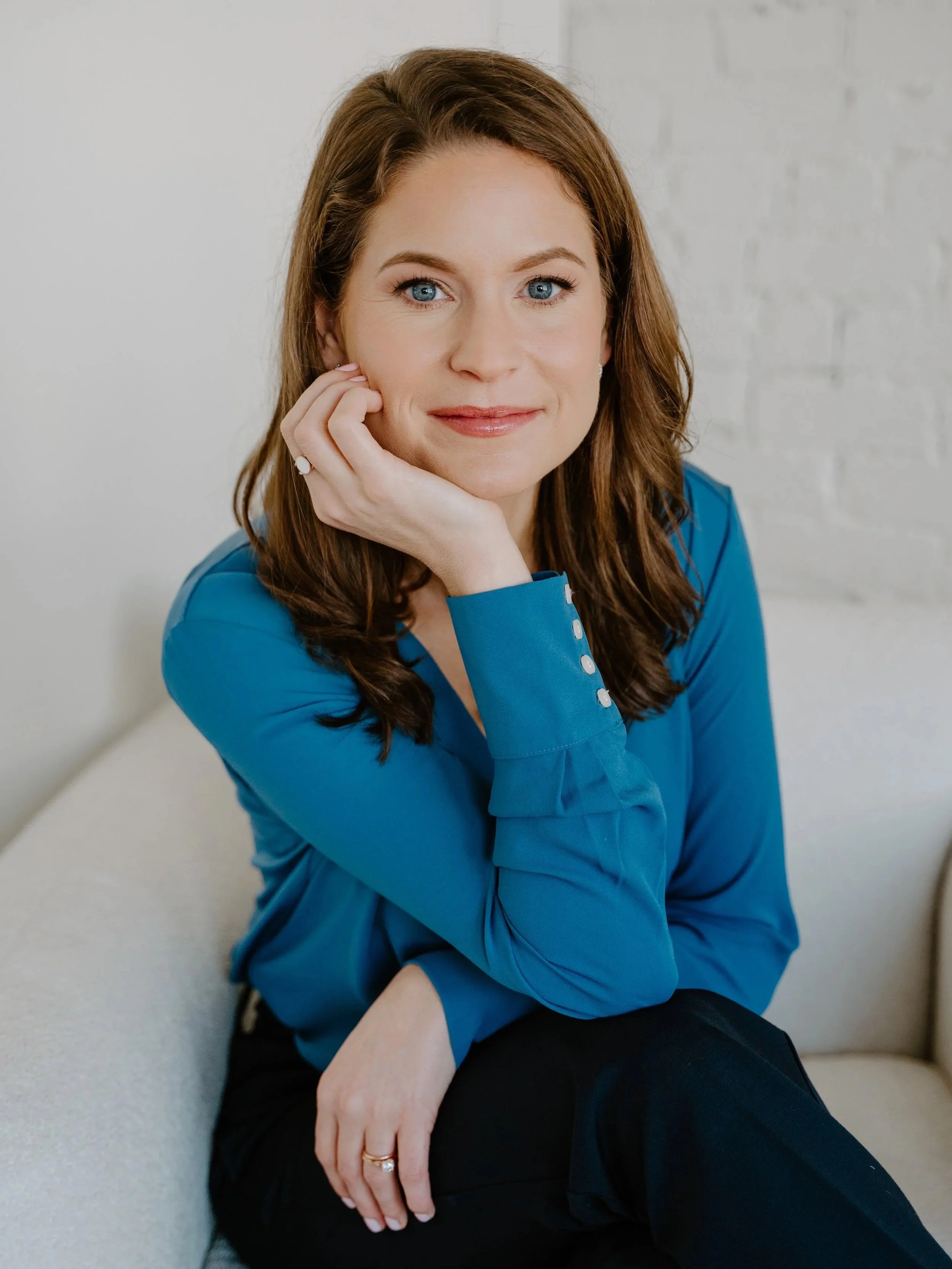 Headshot of a woman with red hair, blue eyes, wearing a blue blouse, sitting on a light-colored sofa, posing with her chin resting on her hand against a white brick wall.