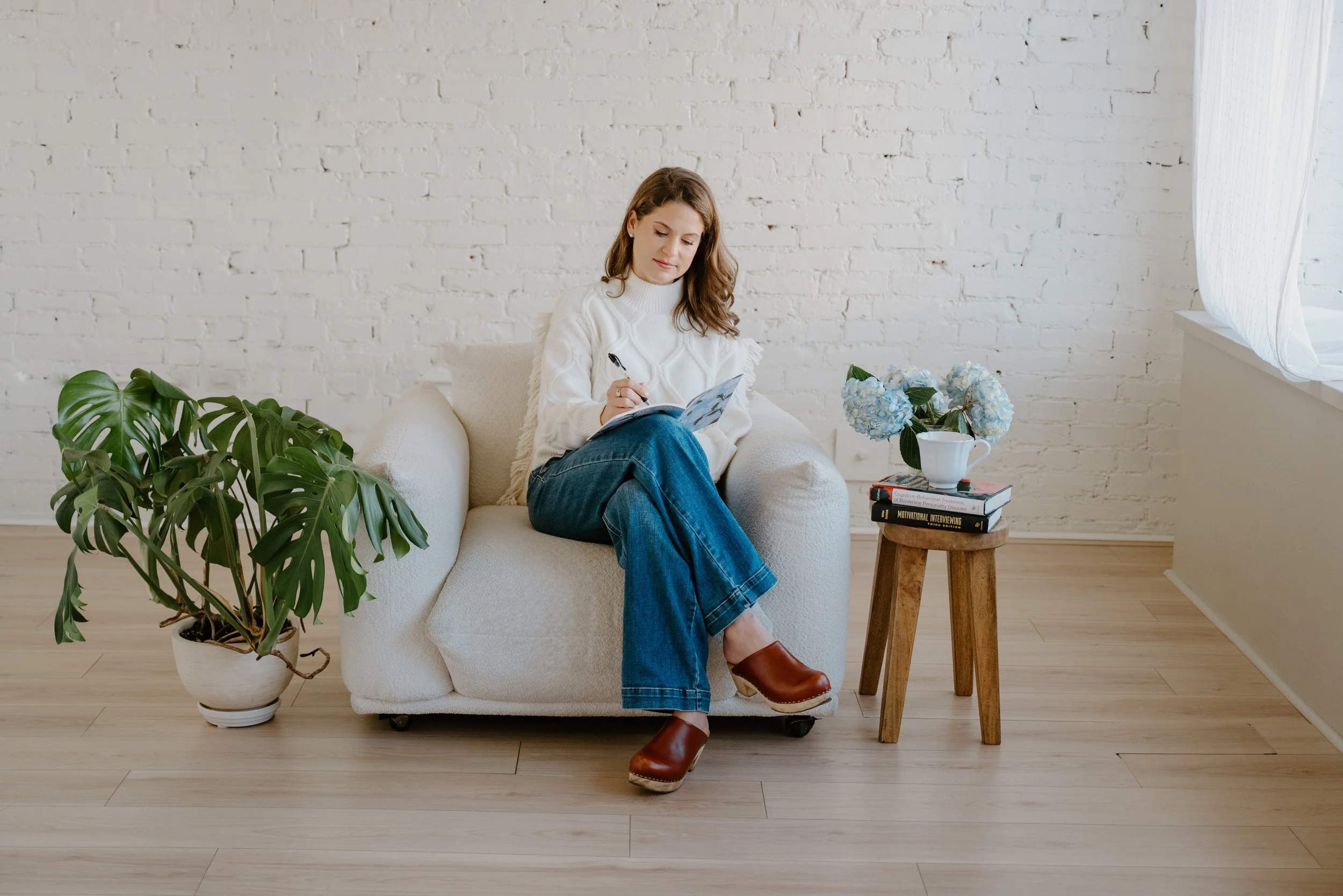 A woman sitting on a white armchair writing in a notebook in a bright room with a white brick wall, a potted plant to her left, a small wooden table with books, a teacup, and a bouquet of blue hydrangeas beside her.
