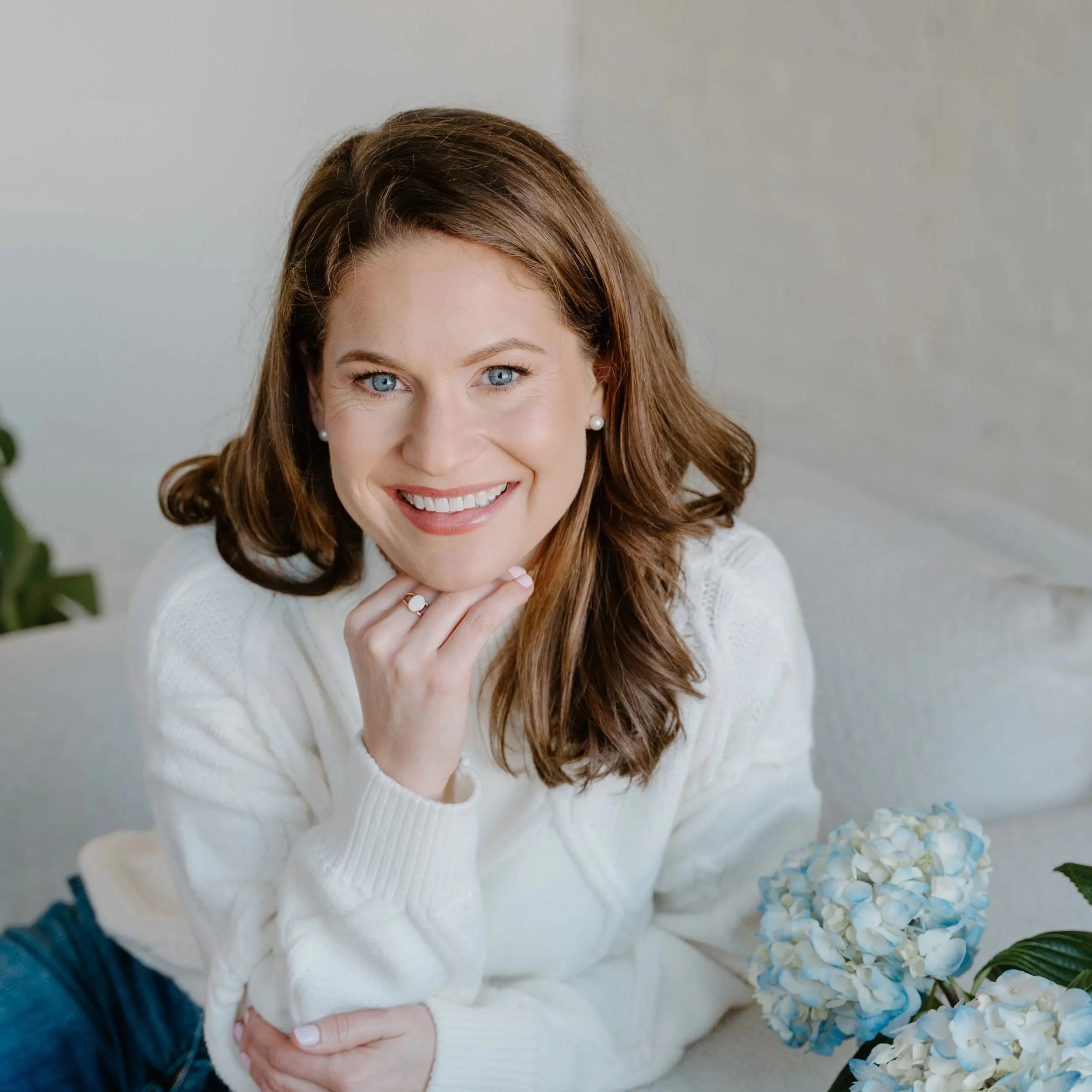 A woman with long red hair and blue eyes smiling, sitting on a white sofa with white and blue hydrangeas nearby.
