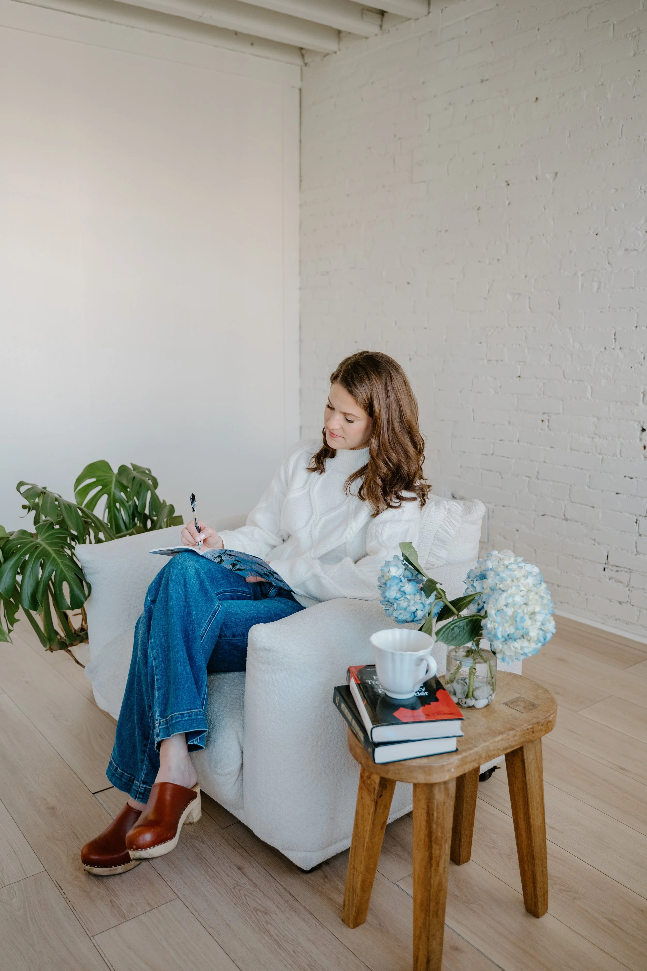 A woman with brown hair sits on a white armchair, writing in a notebook. She wears a white sweater, blue jeans, and brown shoes. Next to her is a small wooden table holding a stack of books, a white cup, and a vase with blue and white hydrangeas. There is a green plant on the floor beside the armchair, and a white brick wall in the background.