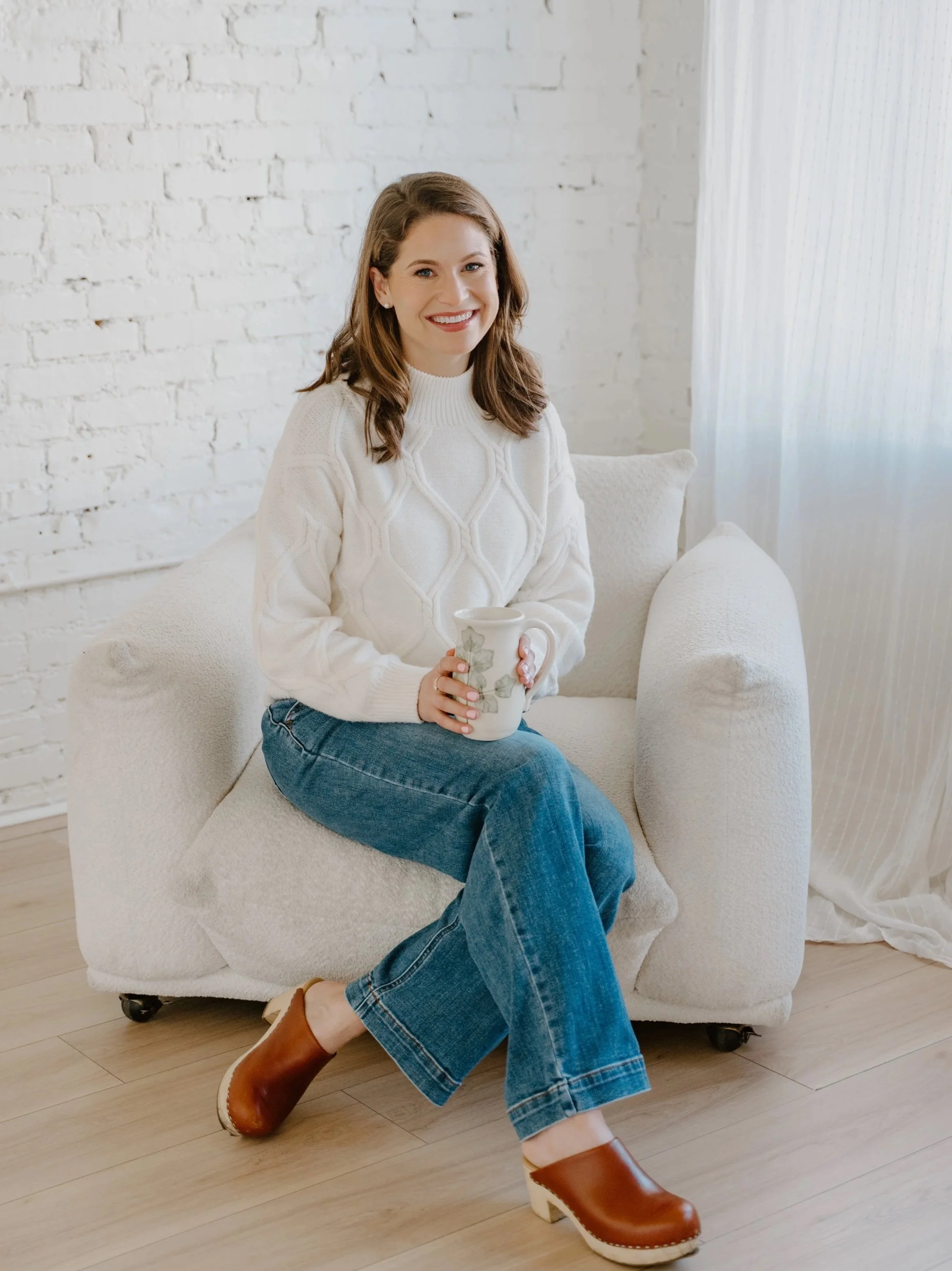 A woman with shoulder-length brown hair sitting on a white sofa, holding a white mug with green leaf design, smiling in a bright room with white brick wall and sheer curtains.