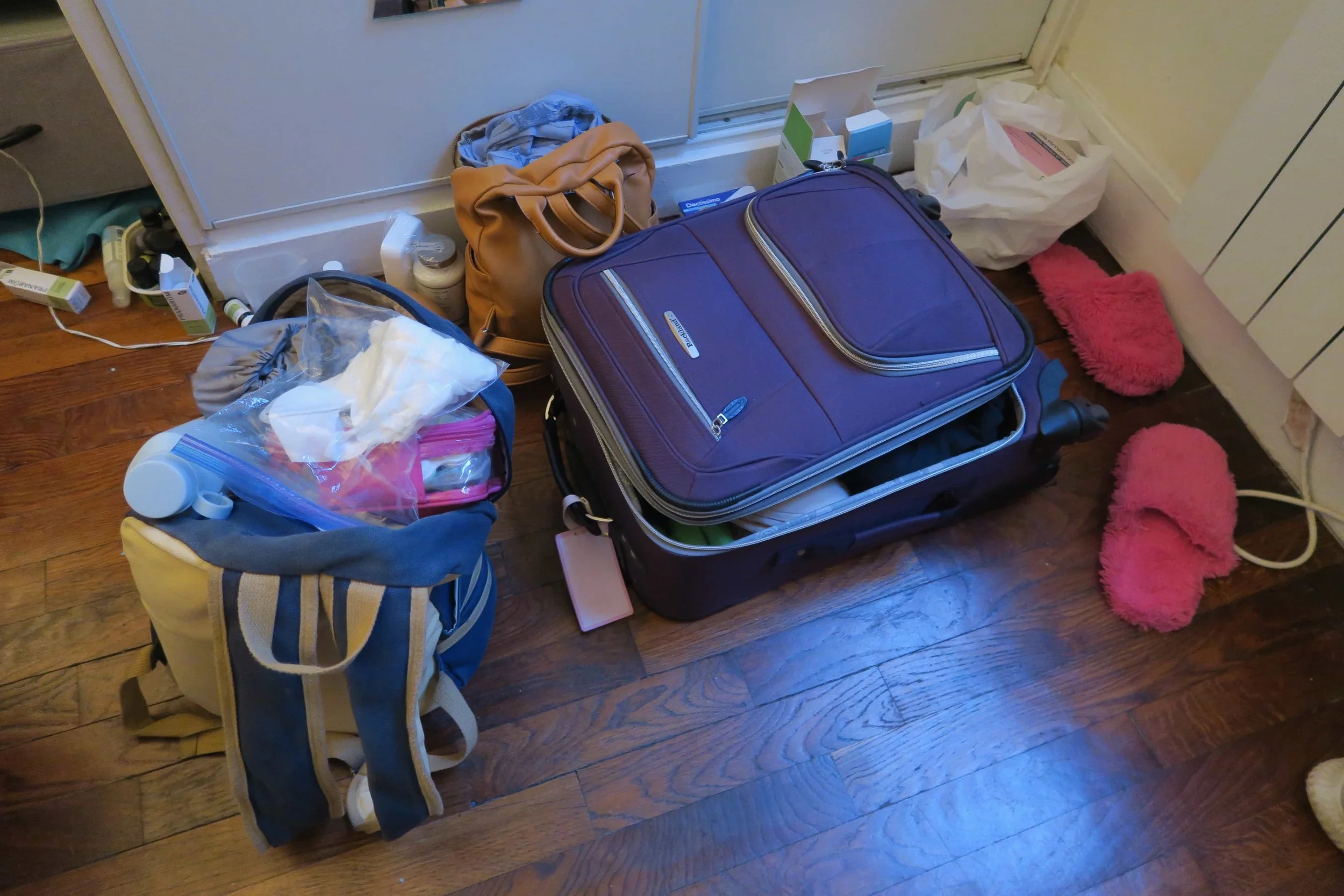 purple suitcase pink slippers tan backpack and blue packpack on hardwood floor