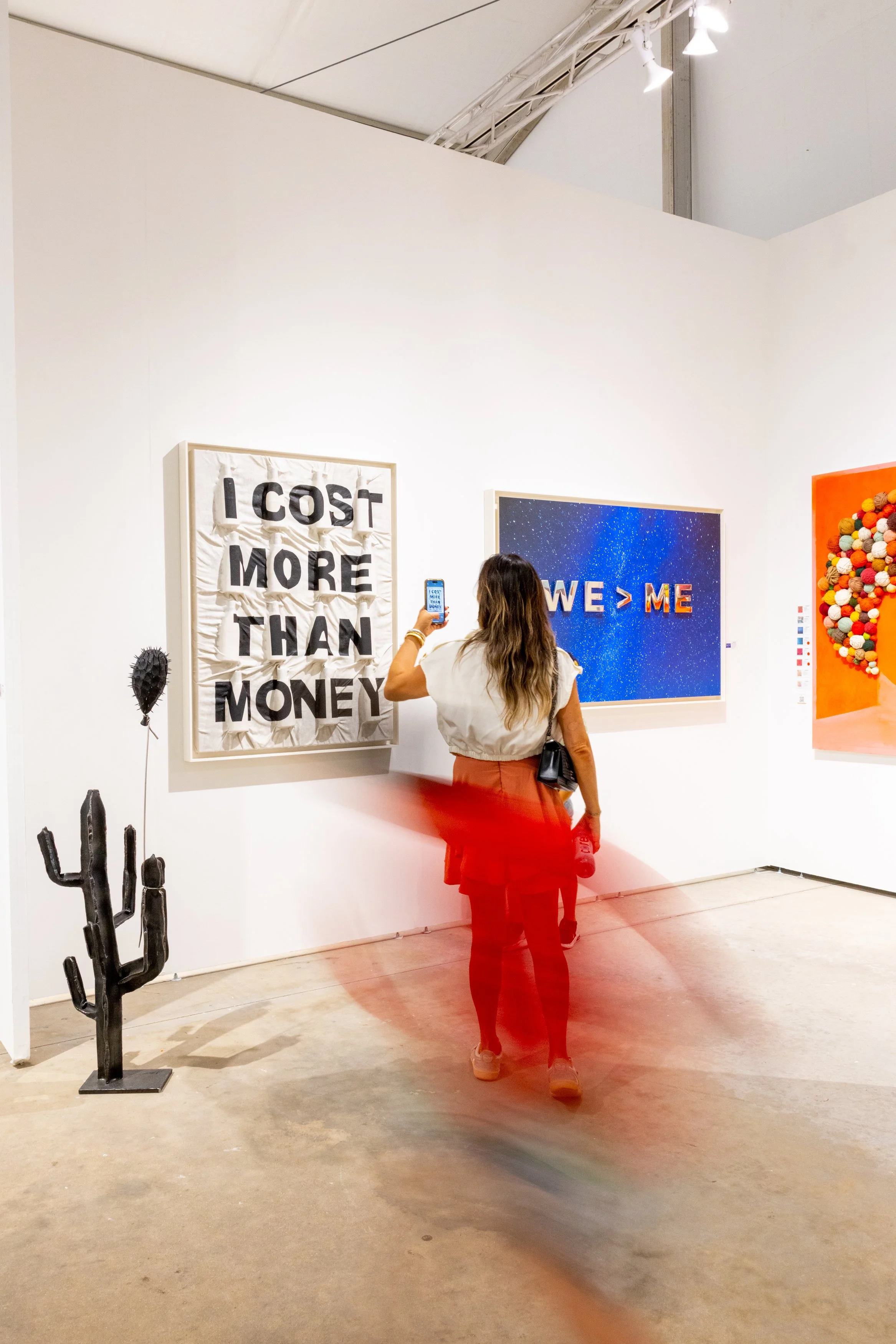 artworks displayed on white wall gallery with audience viewer making a photograph using smart phone and a motion blur.  each of the 2 artworks say, "I COST MORE THAN MONEY" and "WE > ME"