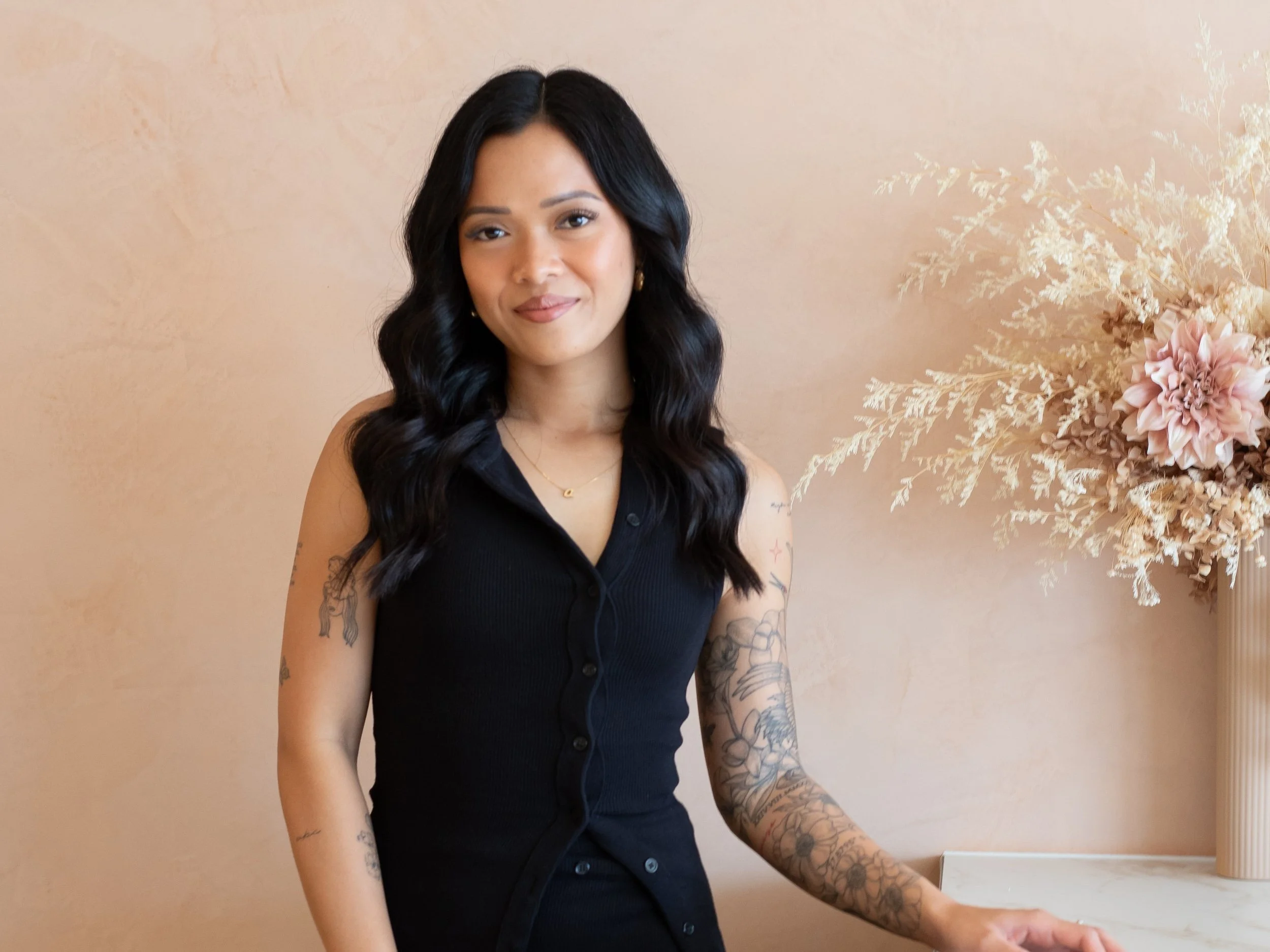 Person with long wavy black hair wearing a black sleeveless dress, standing in front of beige background with floral tattoos on left arm and dried floral arrangement to the side.