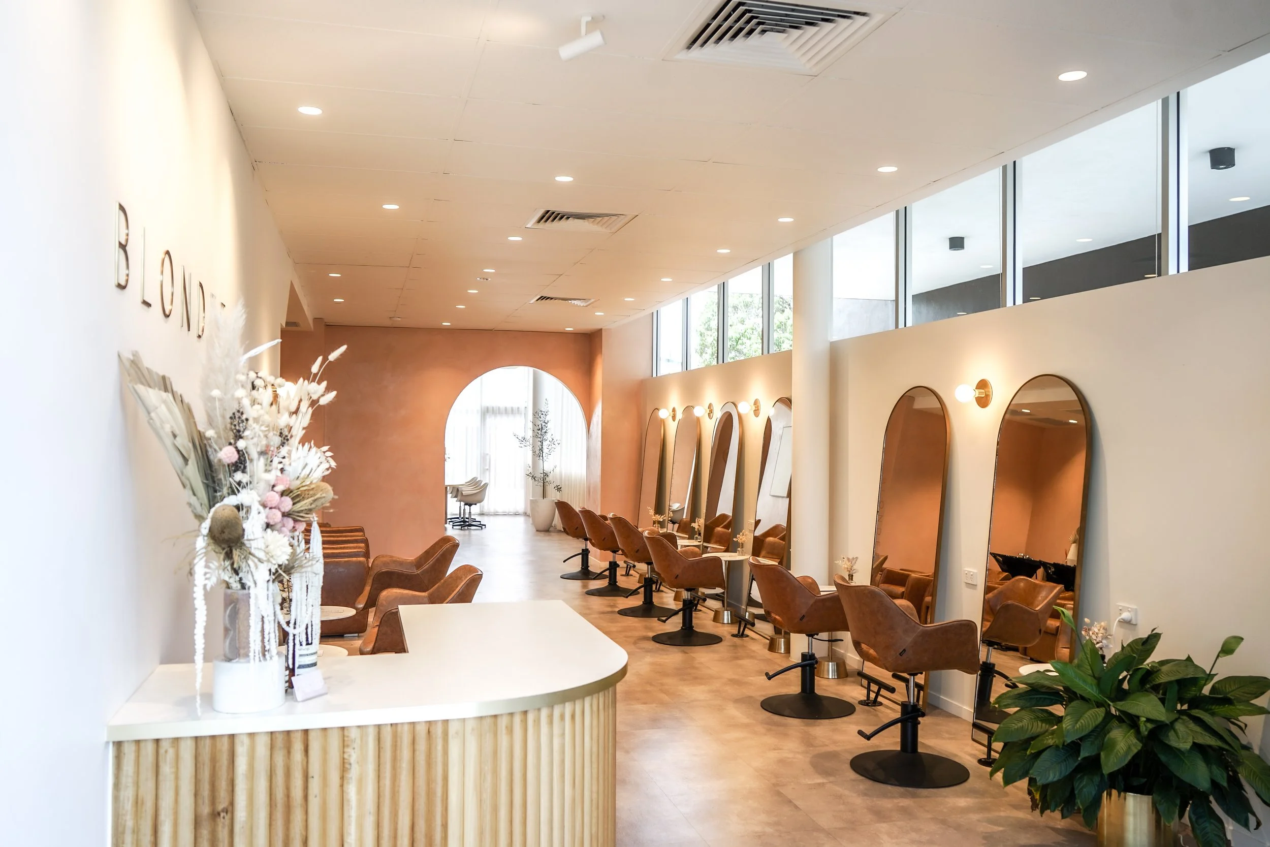 Interior of a modern hair salon with a row of styling chairs and mirrors, a reception desk with decorative flowers, and warm lighting.