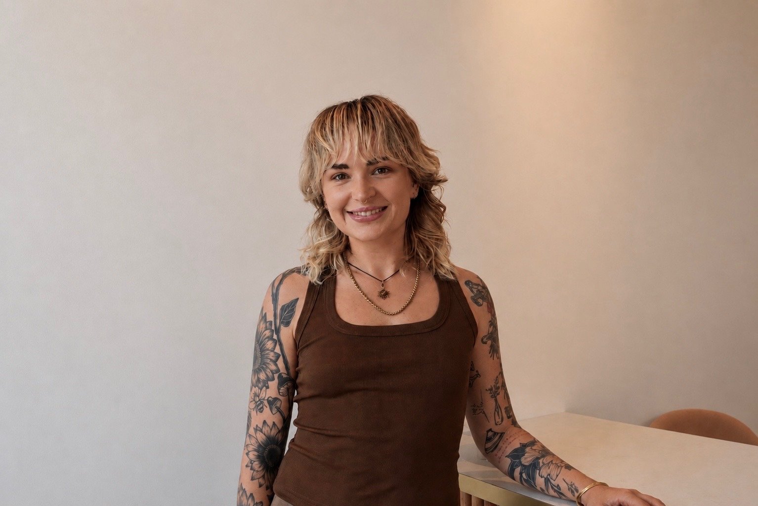 A young woman with blonde hair and tattoos on her arms standing in front of awhite wall with her hand on a desk