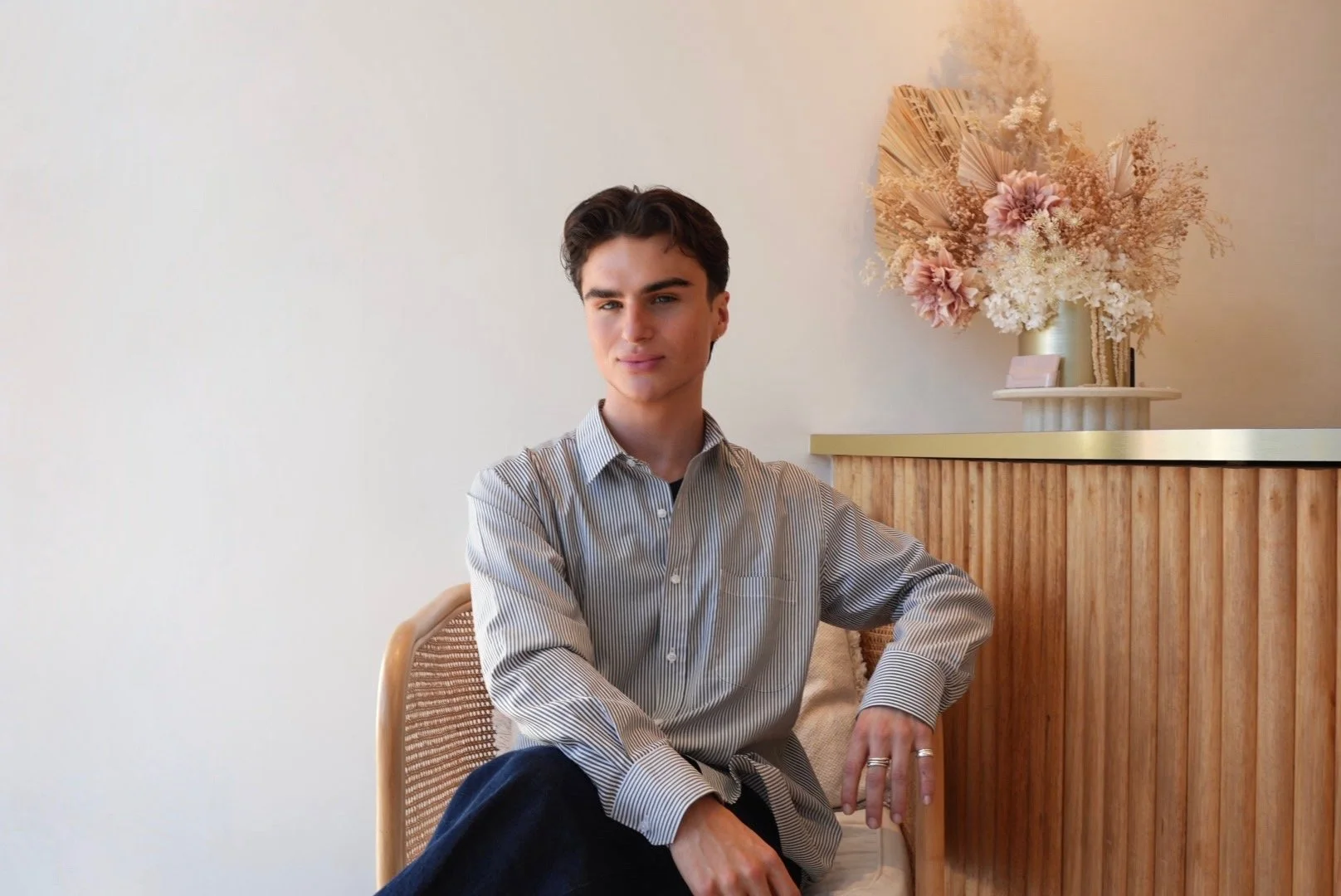 A man with short, dark hair wearing a blue and white striped button up shirt and dark pants sitting in a wicker chair  in front of a reception desk and flowers.