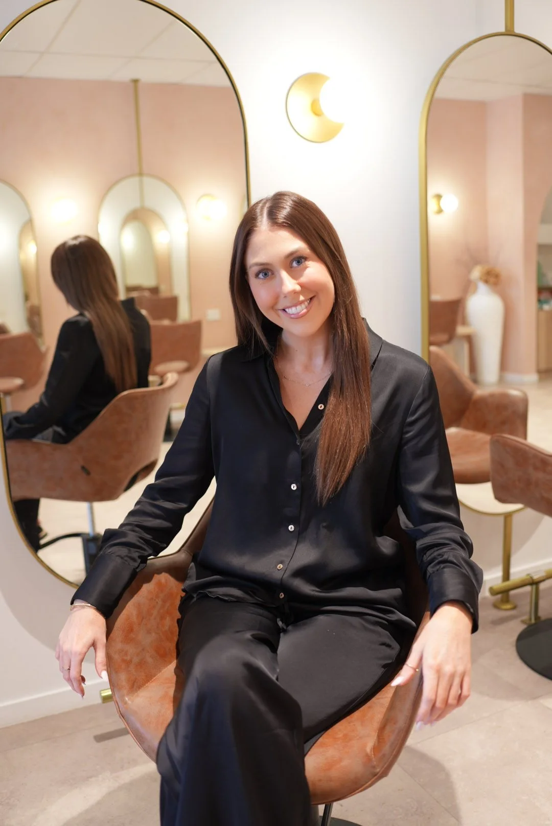 A woman with dark, straight hair and a black silk button up top and pant set sits in a salon chair smiling at the camera. A salon is seen in the background.