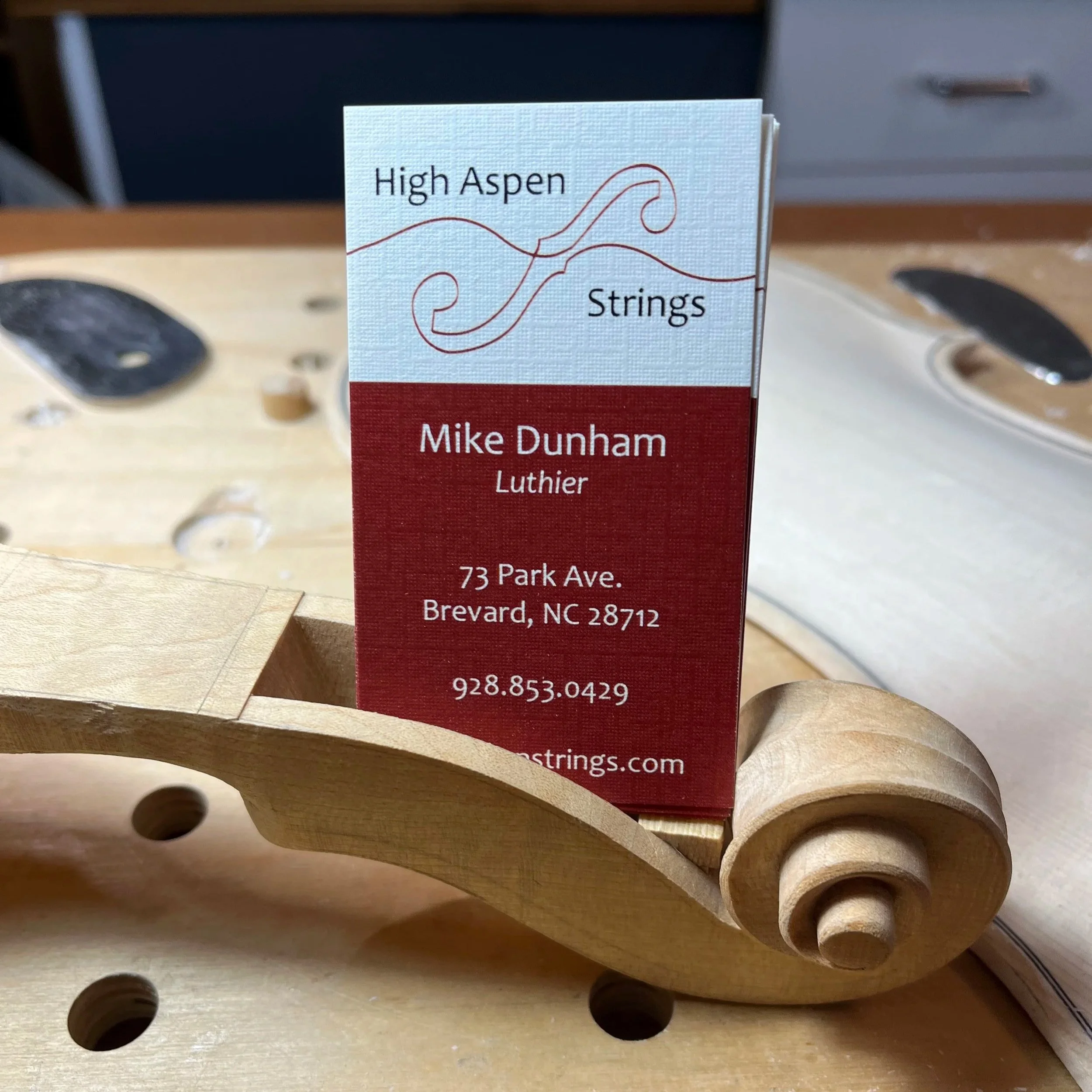 Business card with the text "High Aspen Strings," "Mike Dunham Luthier," address 73 Park Ave., Brevard, NC 28712, phone number 928.853.0429, and website strings.com, placed on a guitar scroll wood piece.