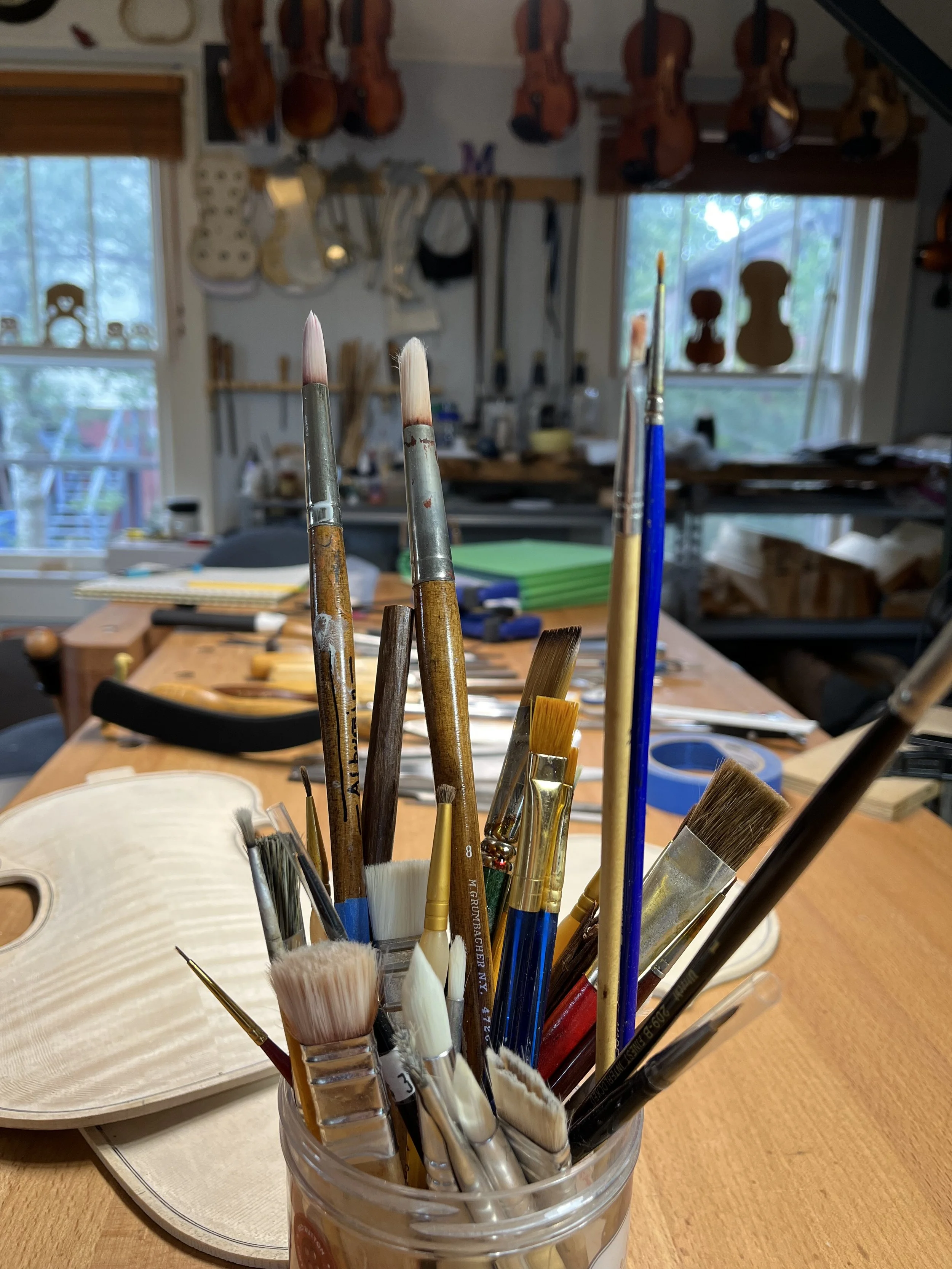 A jar holding various paintbrushes , in a studio with violins hanging on the wall and windows showing trees outside.