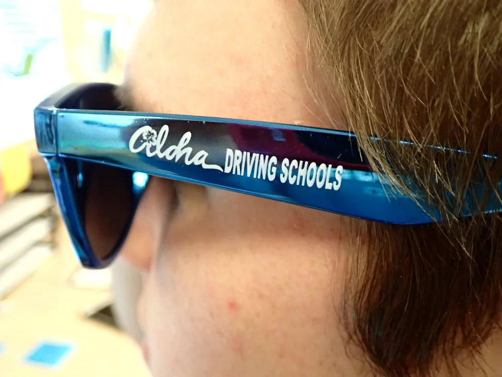 Close-up of a person's face wearing blue sunglasses with the inscription 'Aloha Driving Schools' on the side.