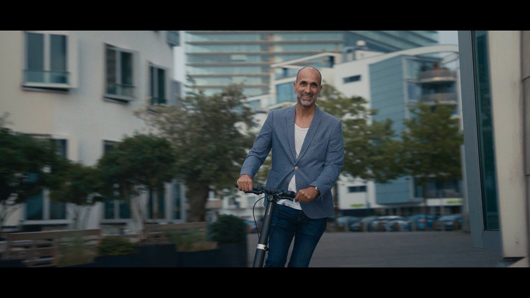 Mann in blauer Jacke fährt mit einem E-Scooter durch eine Stadt, umgeben von modernen Gebäuden.