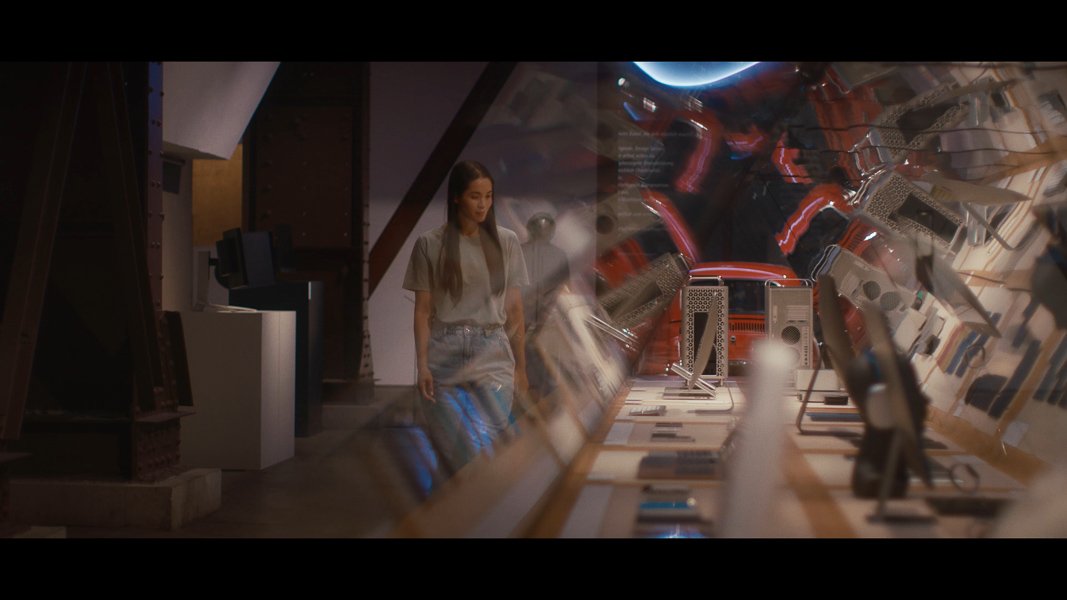 Innenraum eines modernen Museumsausstellungsraums mit einer Vielzahl von Computern und technologischem Equipment auf einem langen Tisch. Eine Person, möglicherweise eine Frau, spaziert durch den Raum.
