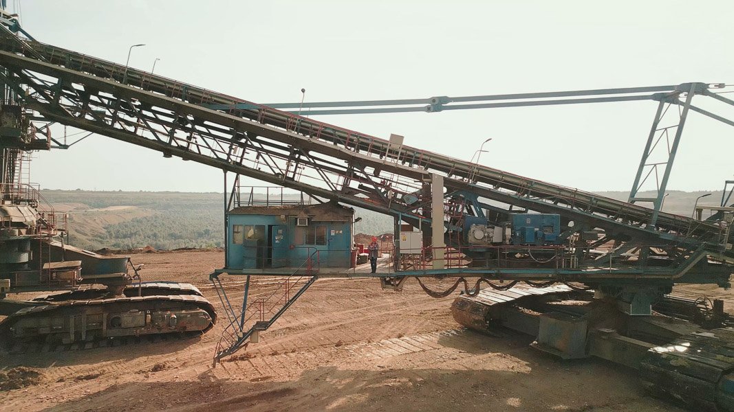 Schweres Bergbaugerät auf einer Baustelle mit Förderband, Kettenfahrwerk und blauem Steuerhaus vor einer hügeligen Landschaft.