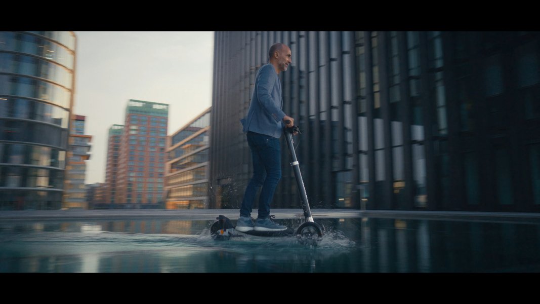 Person auf einem E-Scooter fährt durch flaches Wasser in einer modernen städtischen Umgebung mit Glasgebäuden.