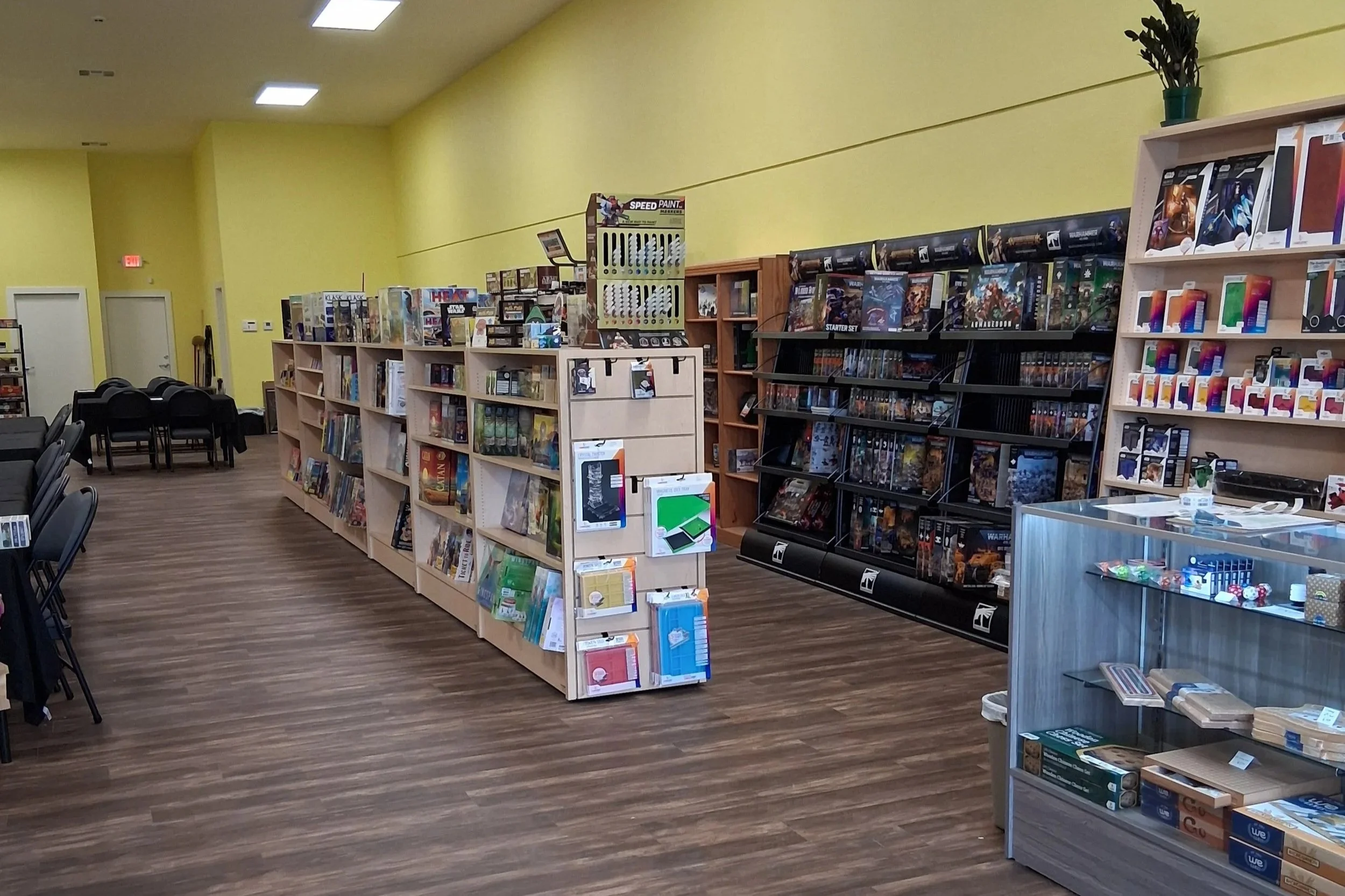Interior of boadgame shop with cheery yellow walls and warm wood floor. Tidy rows of games stretch towards the back where tables and chairs await players.