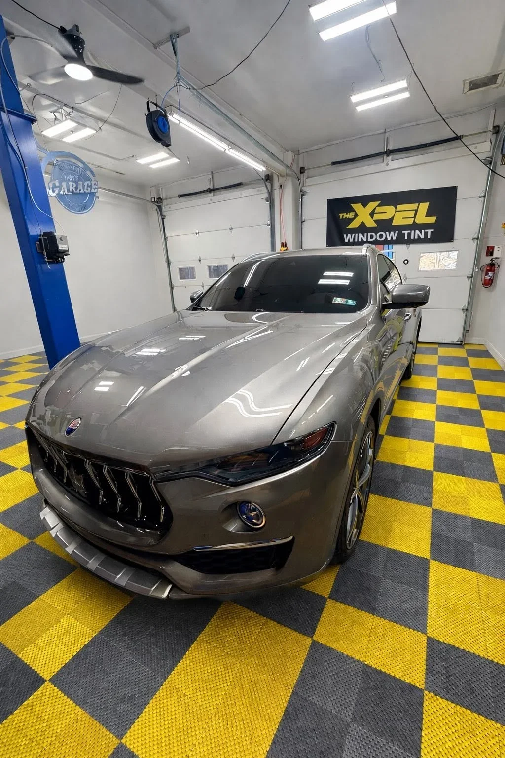 A gray car parked inside a garage with yellow and black checkered flooring. The garage has a white ceiling with bright lights, a blue lift, a ceiling fan, and a black and yellow sign that says 'The Xpel Window Tint'.
