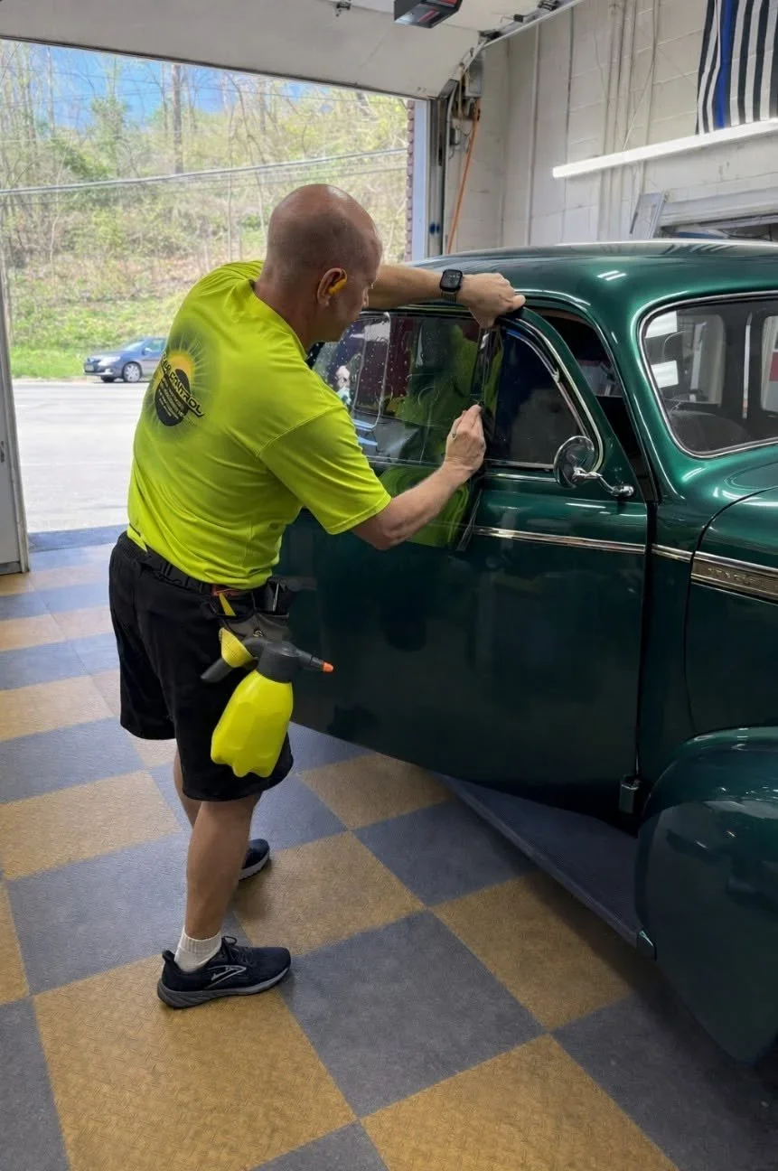 A man cleaning a vintage green car inside a garage, wearing a yellow shirt, black shorts, and black sneakers, with a yellow spray bottle attached to his waist.