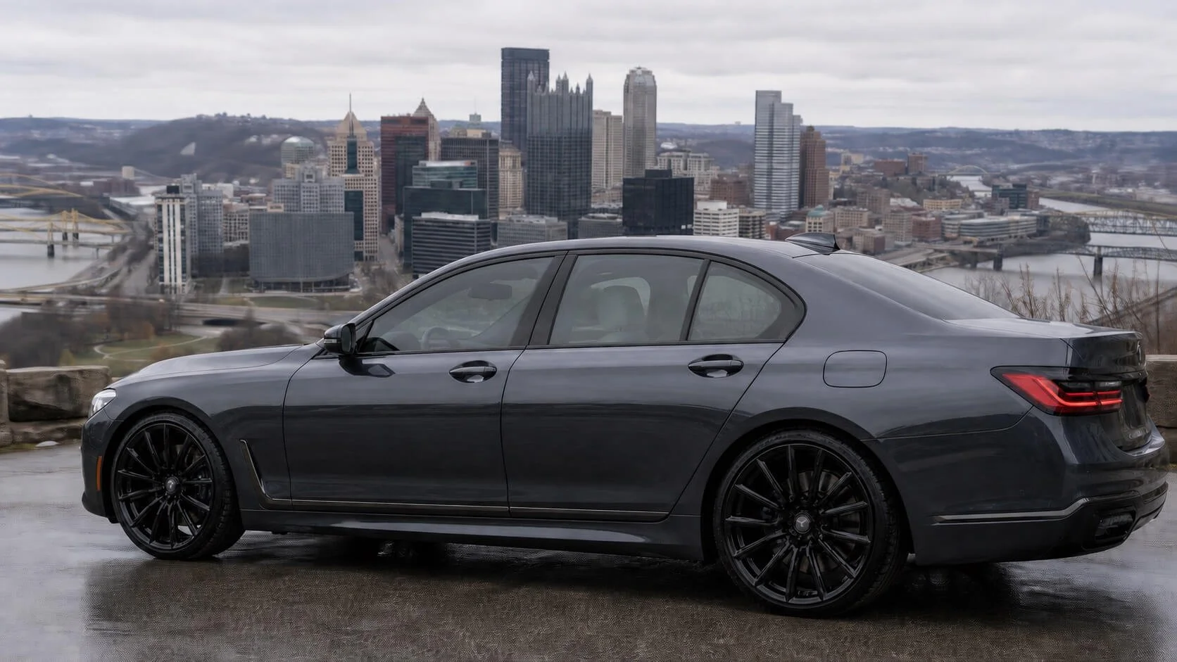 A black luxury sedan parked on a scenic overlook with a city skyline in the background, featuring tall skyscrapers and a river.