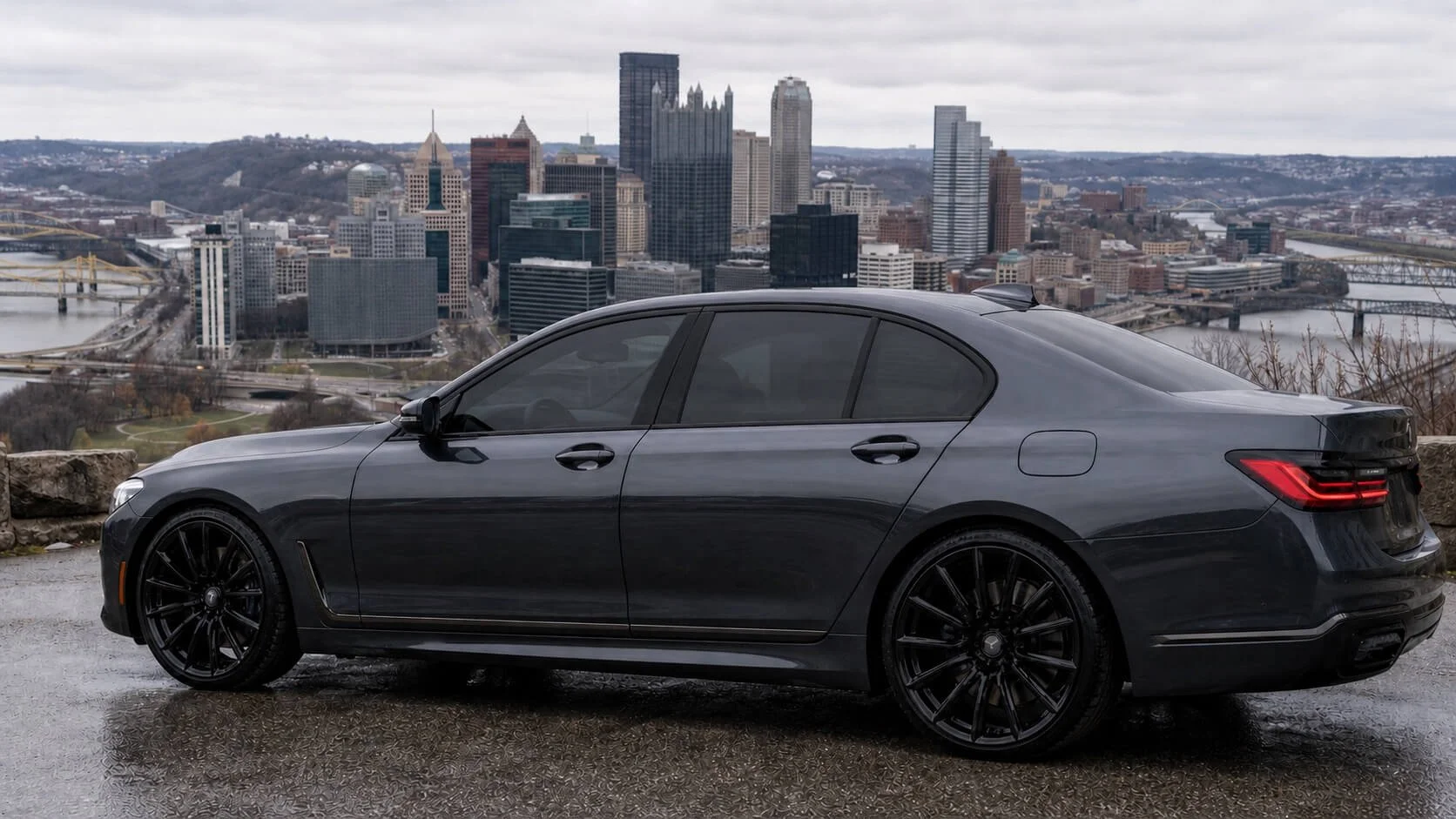 Black luxury sedan parked on a wet road with a city skyline in the background, featuring tall buildings, bridges, and a river under an overcast sky.