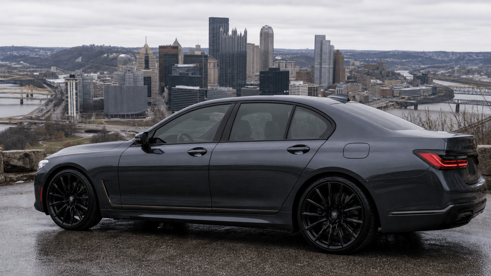 Black luxury sedan car parked on a wet road with a city skyline in the background, including tall buildings, bridges, and rivers under a cloudy sky.