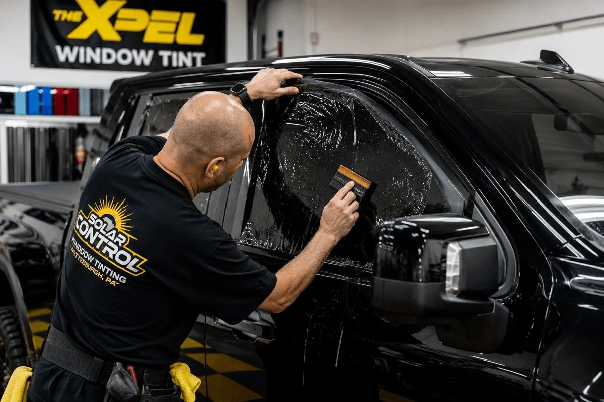 A man in a black shirt with yellow text and logo working on a black vehicle in a workshop. The man is applying tint film to the vehicle's window with a squeegee, and the window has the film partially applied. The background features a banner and various equipment.