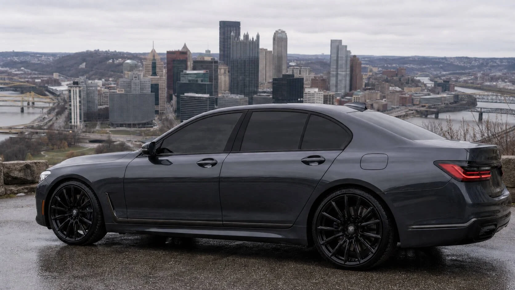 A black luxury sedan with tinted windows parked on a scenic overlook with a city skyline in the background, featuring high-rise buildings, bridges, and a river under an overcast sky.