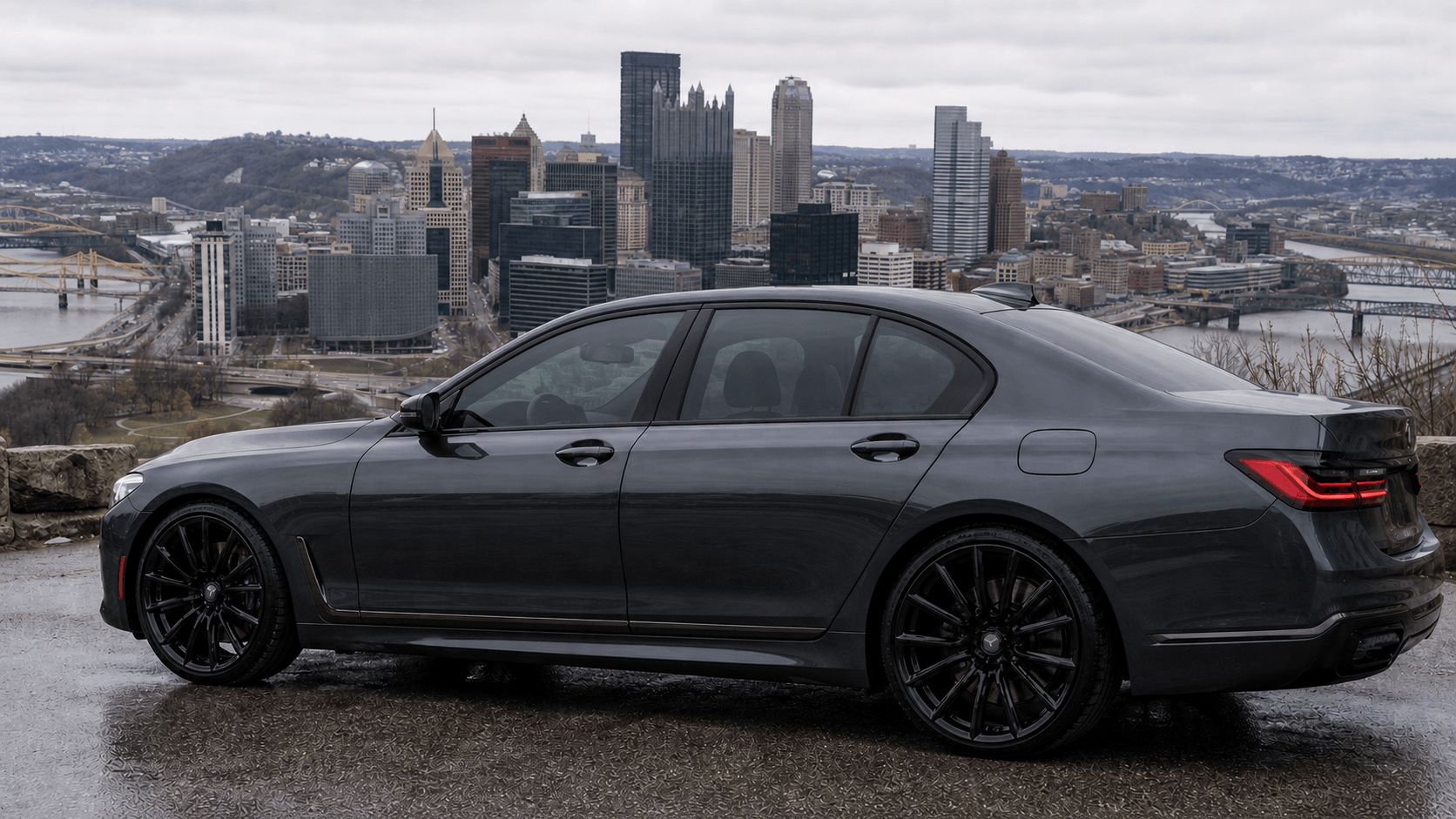 A black sedan parked on a wet surface with a city skyline in the background on an overcast day.