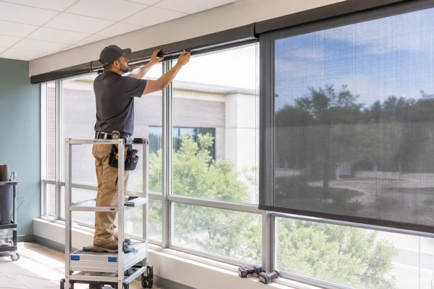 Professional installer mounting commercial solar roller shades above office windows.