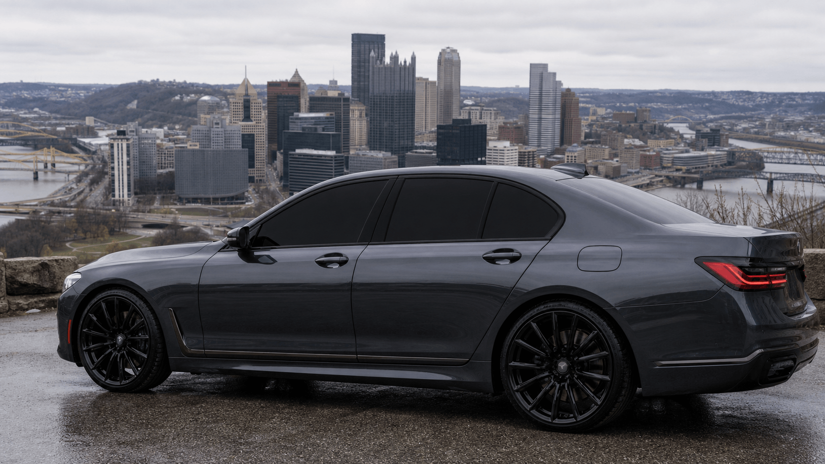A black luxury sedan parked on a wet road with a city skyline and river in the background under an overcast sky.