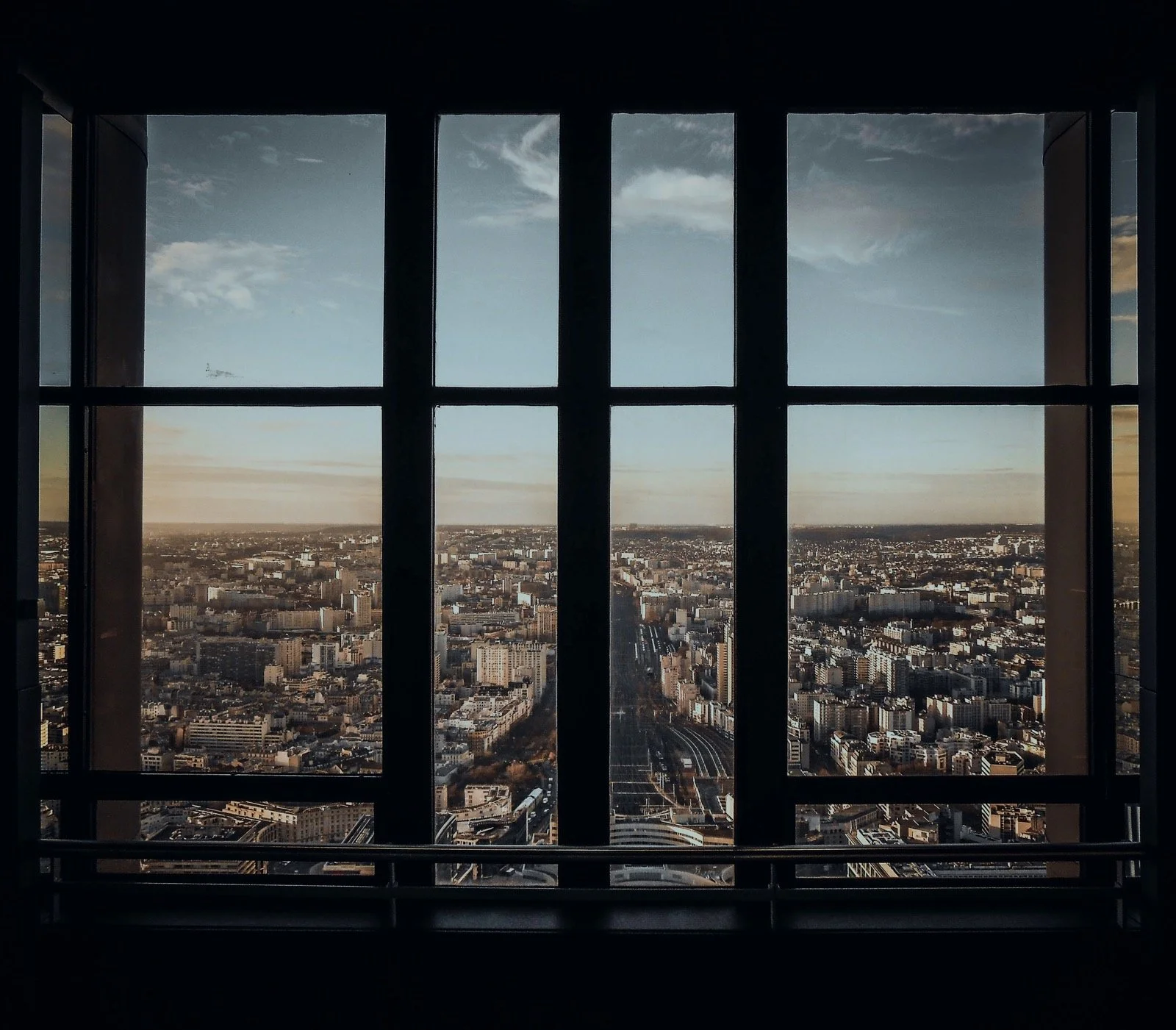 View of a city skyline through a window with vertical and horizontal bars dividing the scene. The city below includes numerous buildings, roads, and some open areas, with a sky featuring clouds.