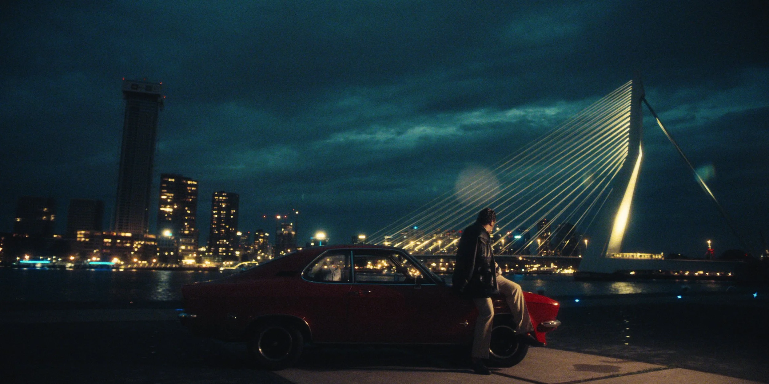 A man sits on the hood of a vintage red car by the river at night, with a modern city skyline and a lit cable-stayed bridge in the background.