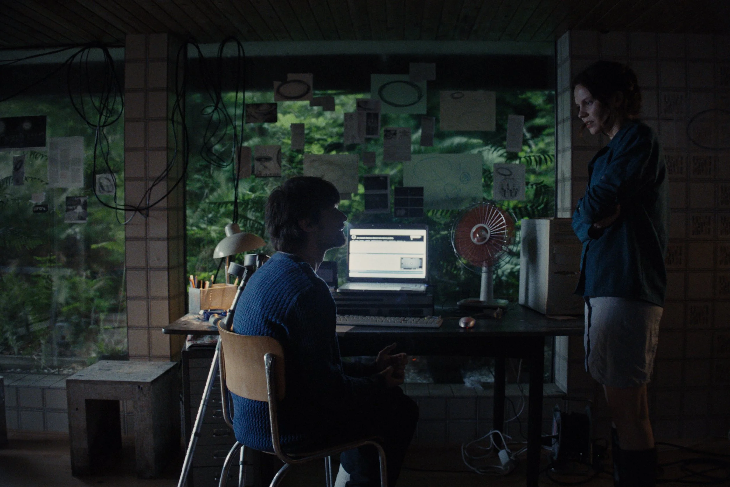 A person sitting at a desk working on a computer, with another person standing and talking in a dimly lit room with a large window showing greenery outside.