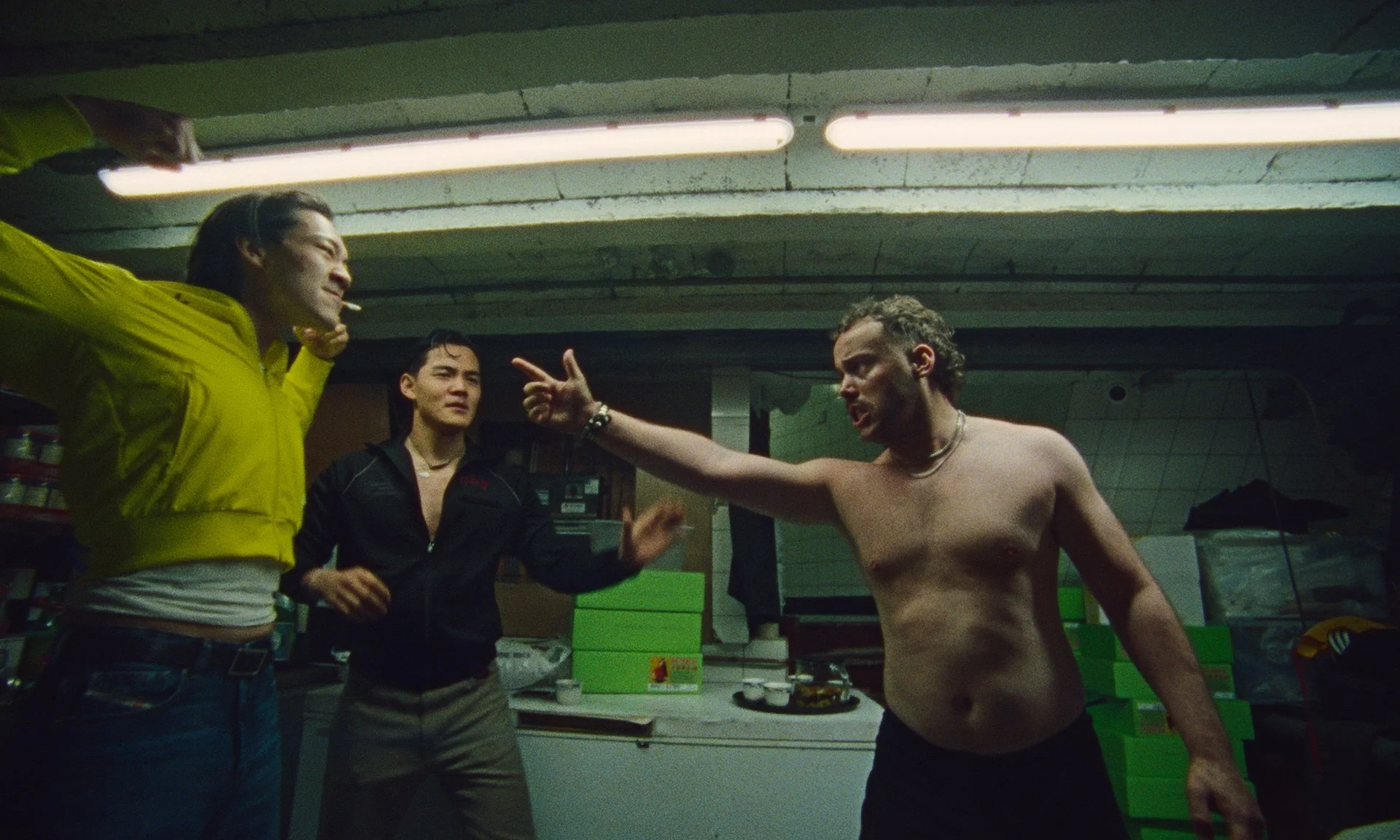 Three people in a heated argument in a dimly lit basement or storage room. One shirtless man with curly hair points aggressively at a woman in a yellow hoodie, while another woman in a black jacket looks on.