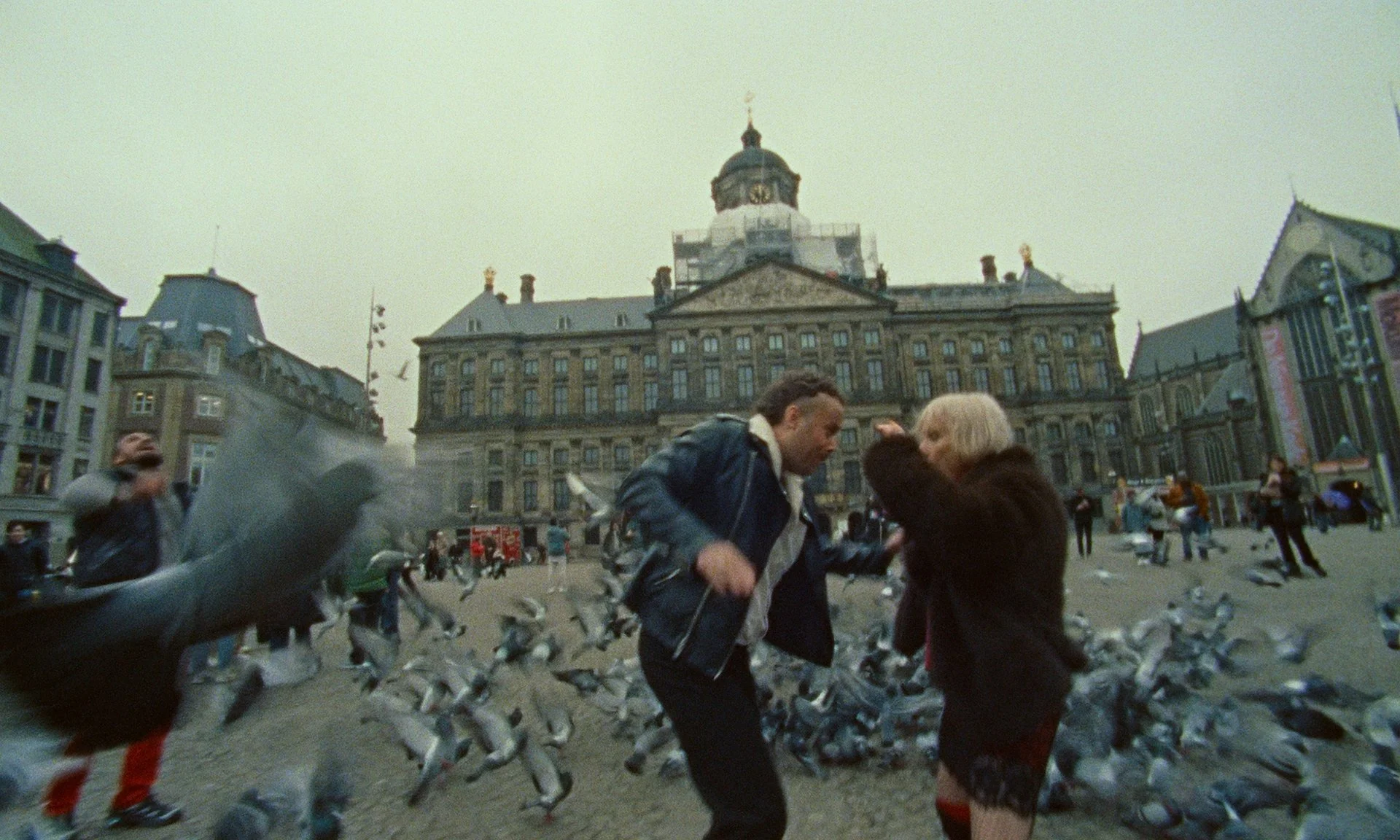 People arguing in a city square surrounded by pigeons, with a historic building in the background.